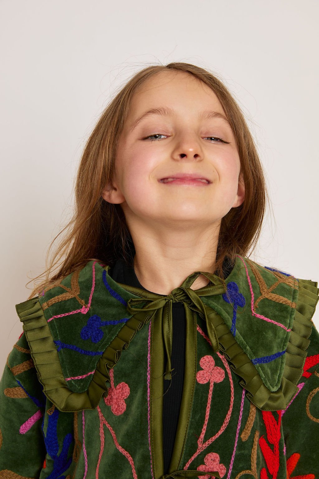 A young girl with long brown hair smiling confidently, wearing a green velvet top with colorful embroidery and ruffled shoulder details.