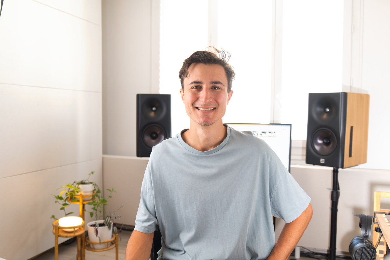 A smiling young man sitting in a music or recording studio surrounded by professional studio monitors and equipment, with a computer screen and some potted plants in the background.