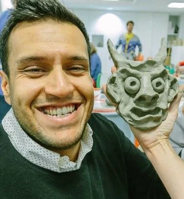 Man smiling and holding a clay sculpture of a face with exaggerated features.