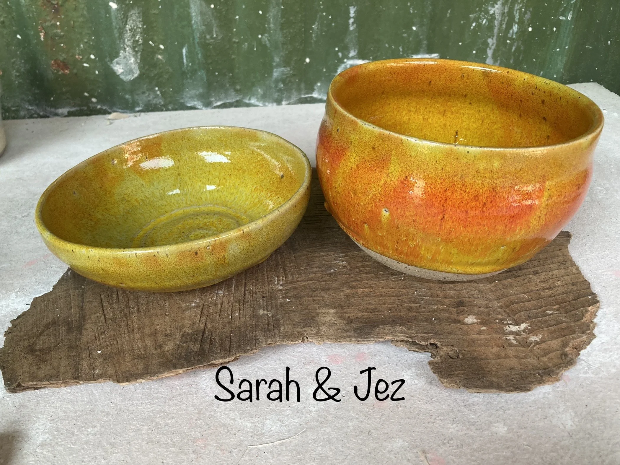 Two ceramic bowls, one smaller and one larger, with yellow and orange glaze, are placed on a piece of rough, weathered wood. The background shows a green wall with some reflections and scratches. There is text in black font at the bottom of the image