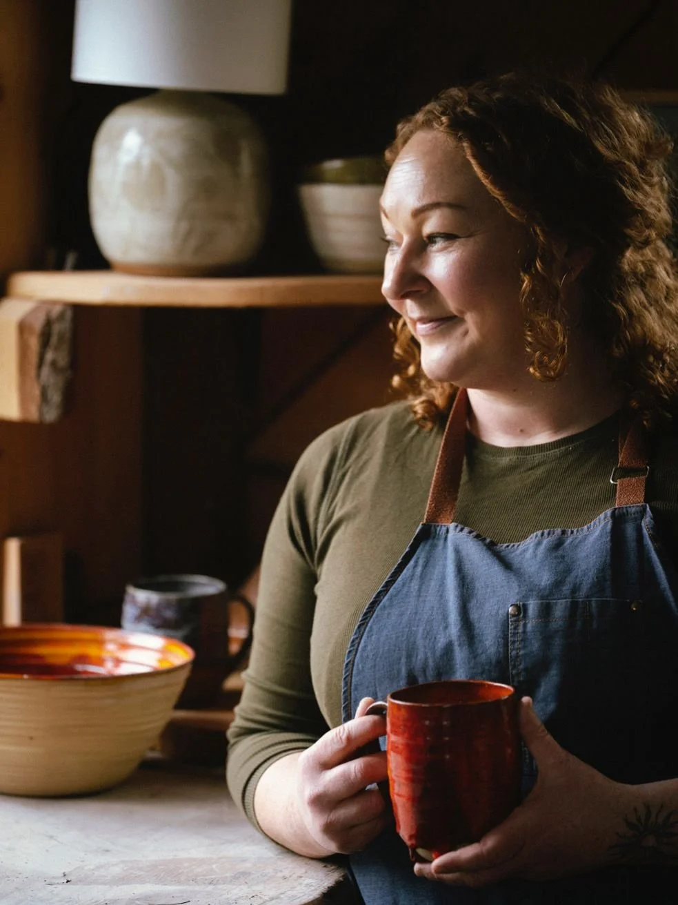 Charlie Clarke in an apron smiles, holding a red mug, with pottery bowls and mugs displayed in the background.