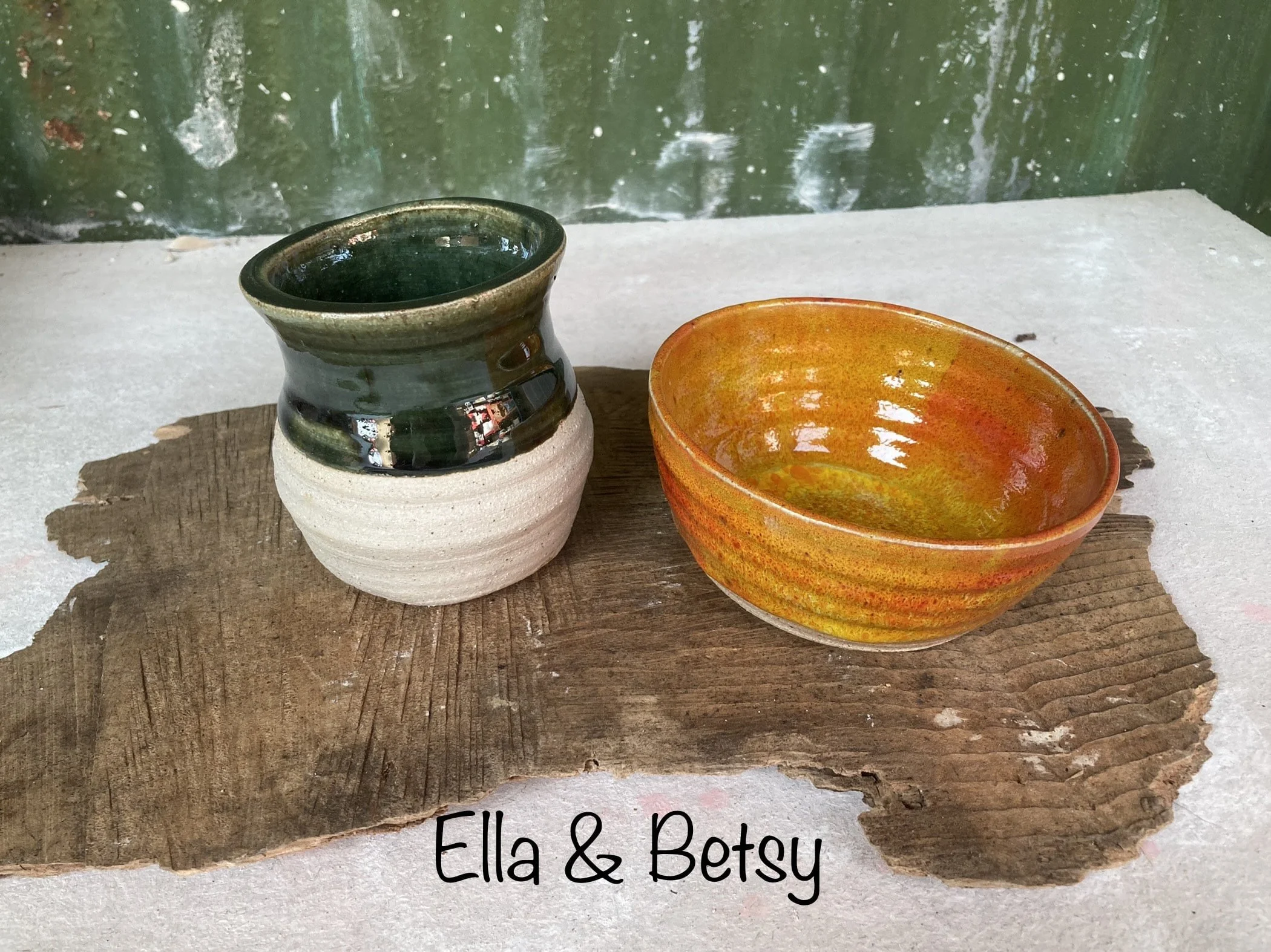 Two ceramic bowls, one green and white and the other orange and yellow, placed on a rustic wooden piece on a grey surface, with a green wall in the background. Text on the image reads 'Ella & Betsy'.