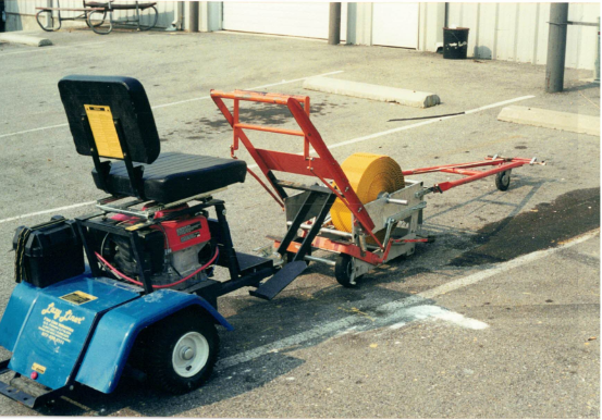 A contrast tape machine with a seat, battery, and tool storage, attached to a large orange and yellow ground power unit on a parking lot