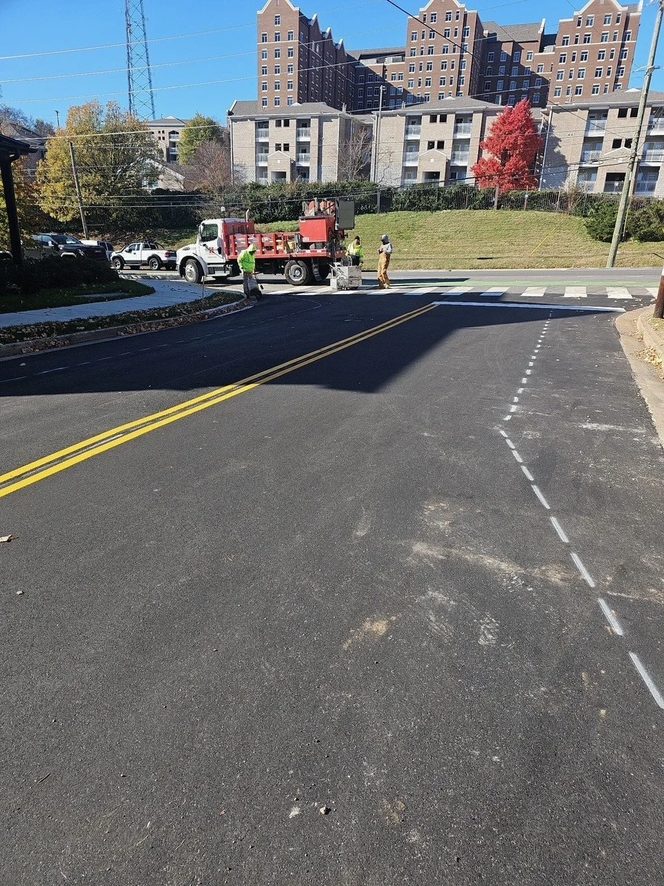 Construction workers working on a crosswalk in a neighborhood with apartment buildings in the background. A fire truck is present, and there are vehicles on the street.