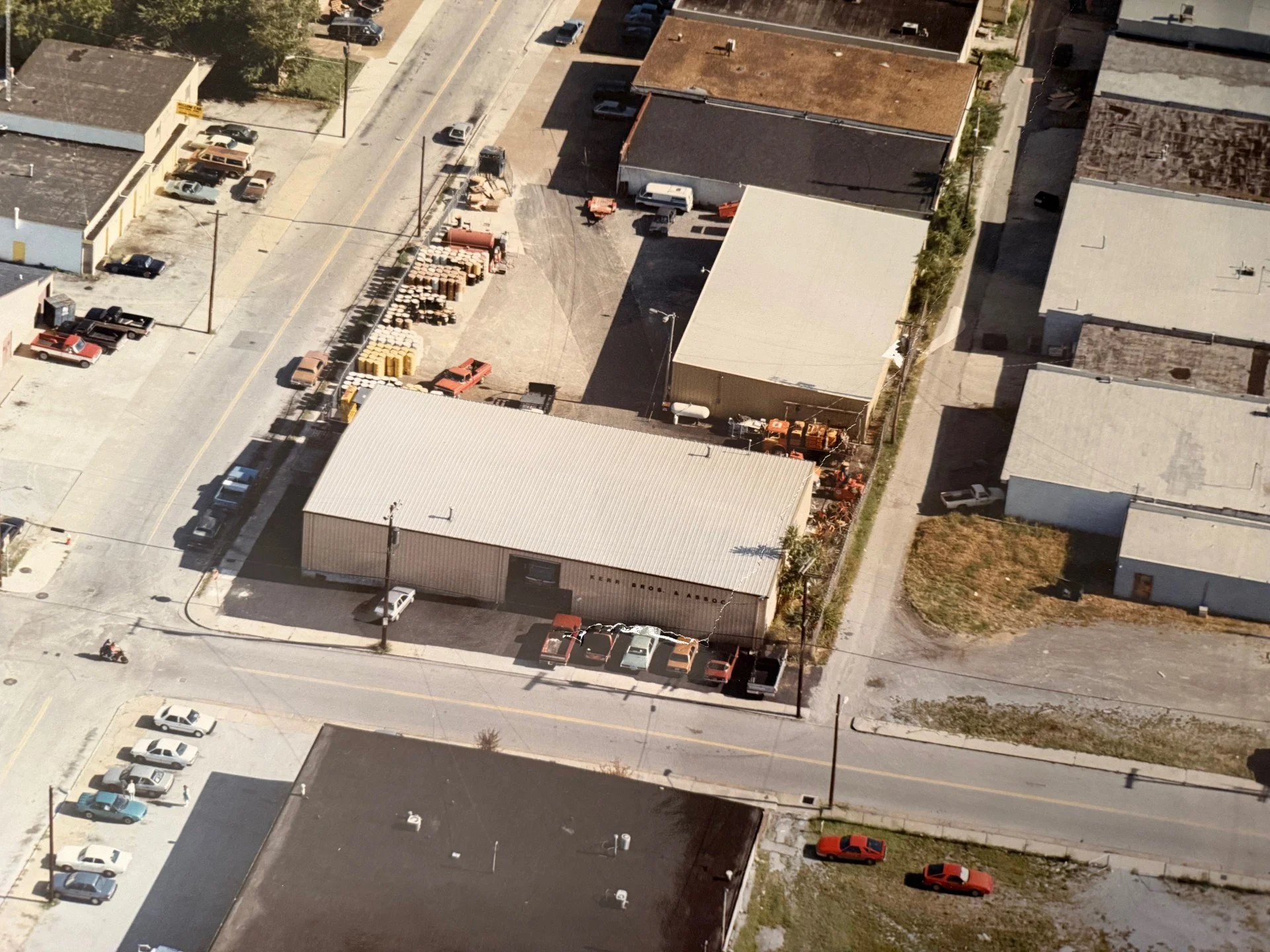 An aerial view of a small commercial area with a warehouse building, a parking lot with several cars, and a few surrounding buildings.