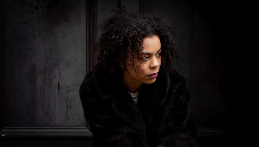 A woman with curly hair, wearing a black coat, sitting against a dark wall, looking to her right with a serious expression.