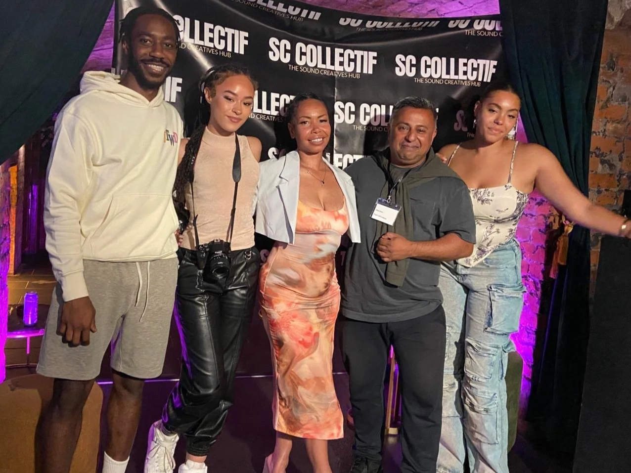 A group of five people posing in front of a black backdrop with 'SC Collectif' written on it. The group includes men and women dressed casually and stylishly, with some wearing athletic wear and others in dresses. They are smiling and standing close together, with some wearing headphones and a camera hanging from one person's neck.