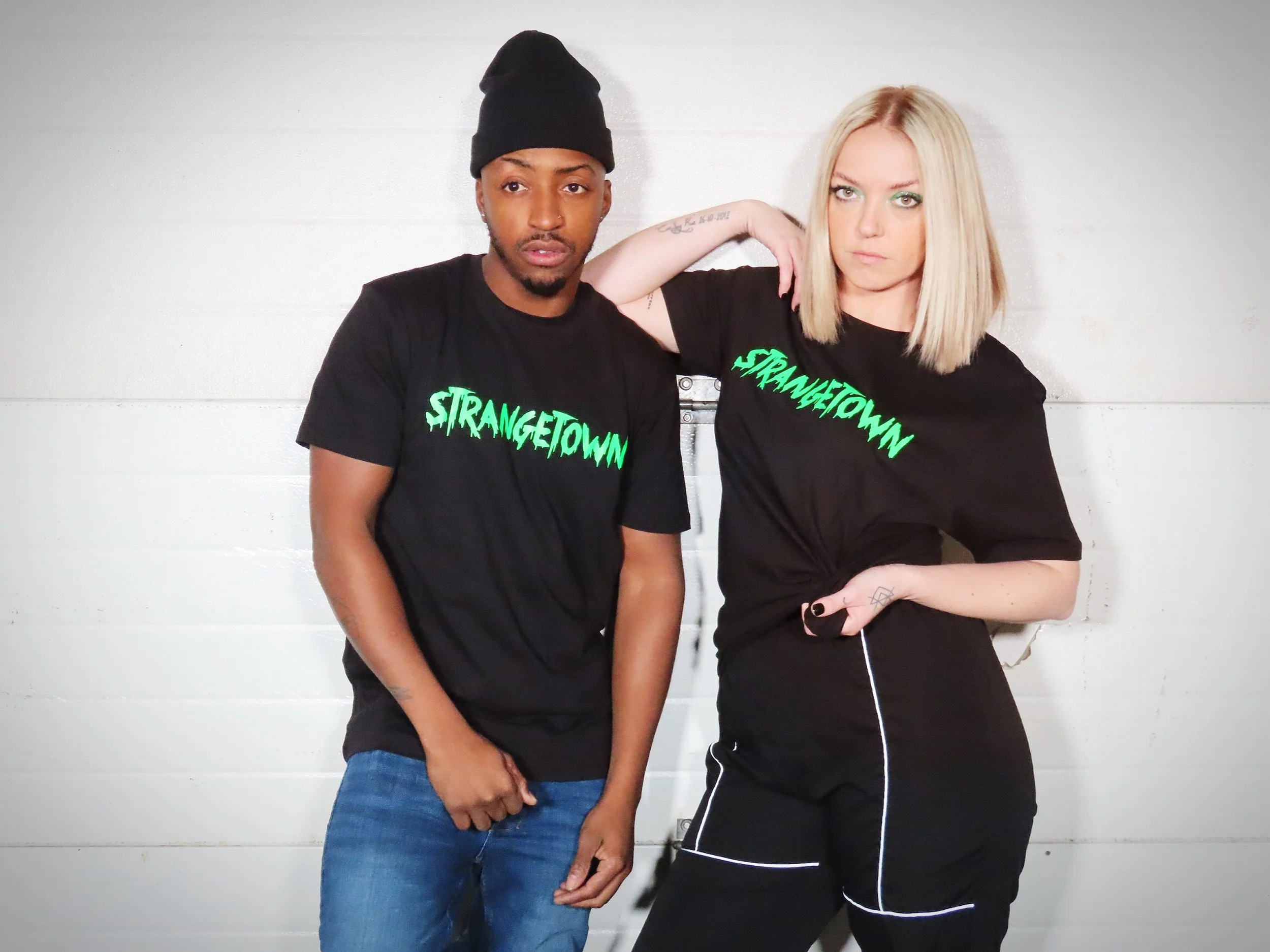 Two young people with serious expressions standing against a white paneled wall, both wearing black T-shirts with 'STRANGETOWN' printed in green.