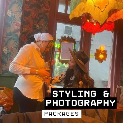 Two women in a colorful, vintage-style room with tropical wallpaper and hanging lamps. One woman, wearing a white blouse and headscarf, appears to be styling or applying makeup to the other woman, who is sitting and wearing a hat. Text overlay reads 'Styling & Photography Packages'.
