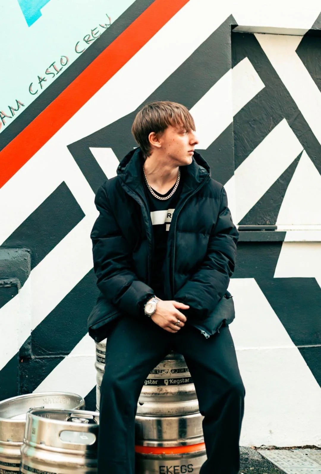 A young man with brown hair sitting on beer kegs in front of a black and white geometric mural, wearing a black jacket, black pants, and accessorized with a chain necklace, watch, and rings.