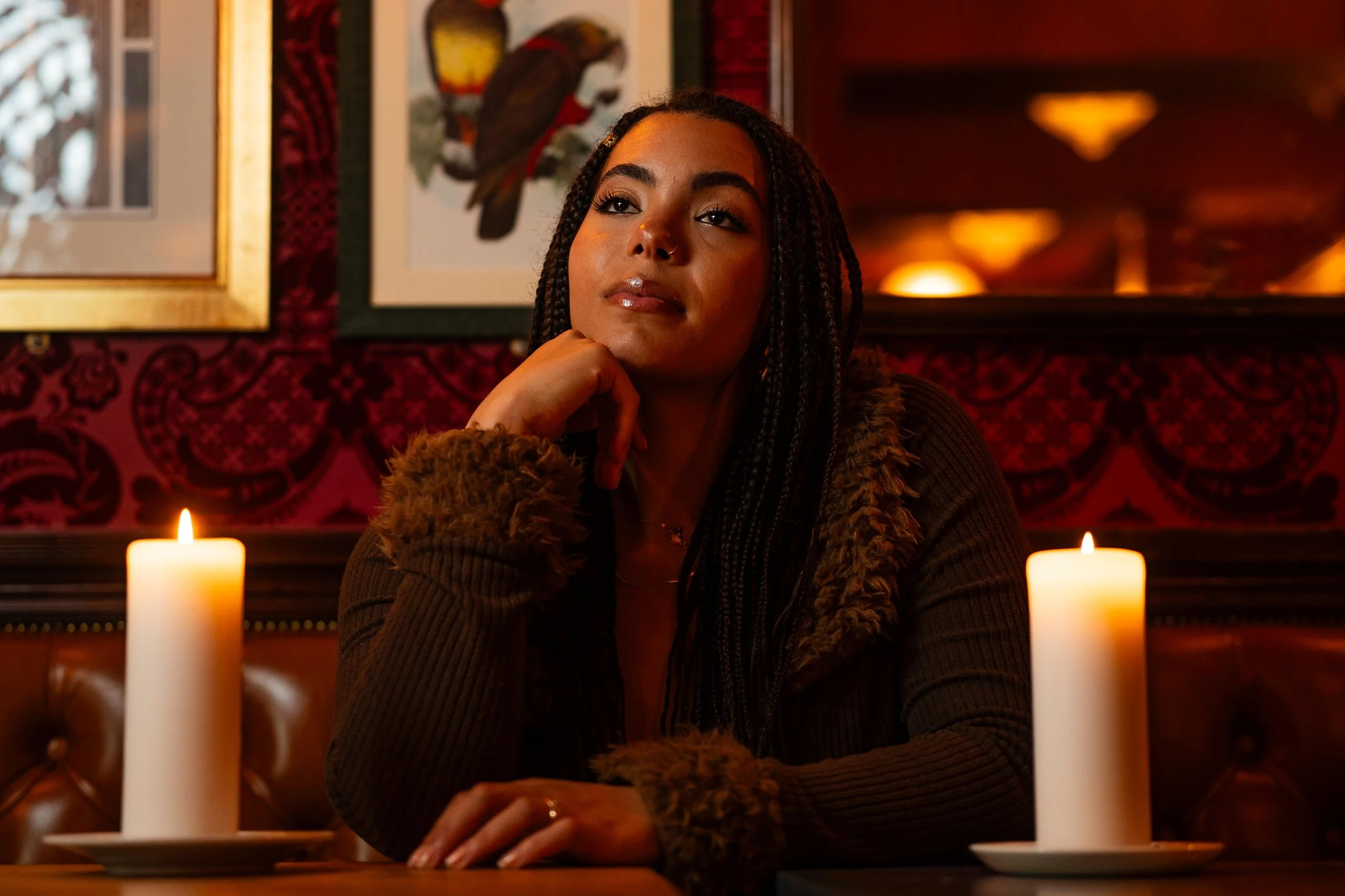 music artist, jessika kay sitting at a restaurant table with her chin resting on her hand, surrounded by lit candles, in a warmly lit, richly decorated interior.