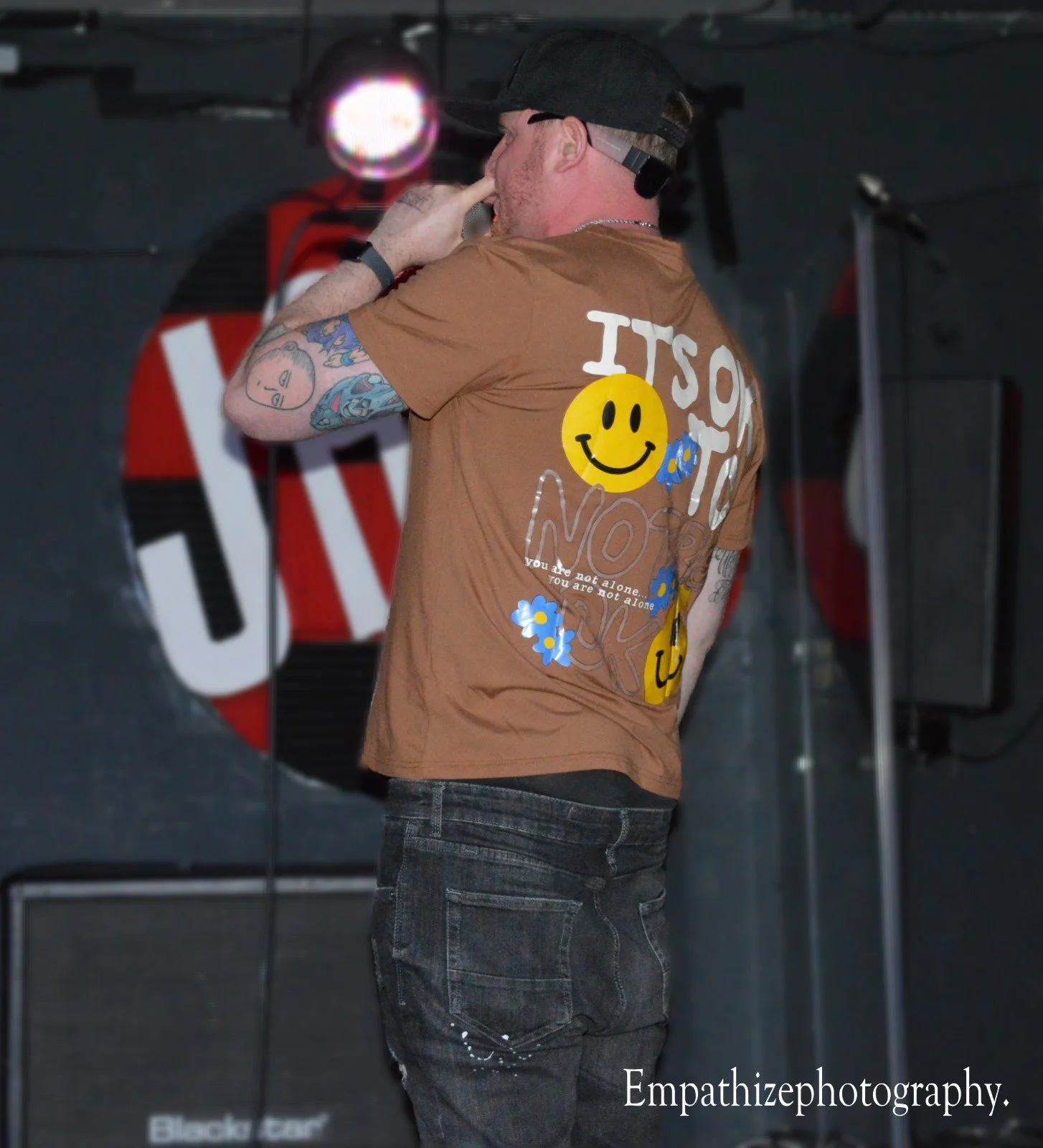A man with tattoos on his arm wearing a brown t-shirt with smiley face and flower graphics, speaking into a microphone on stage with a dark background and musical equipment.