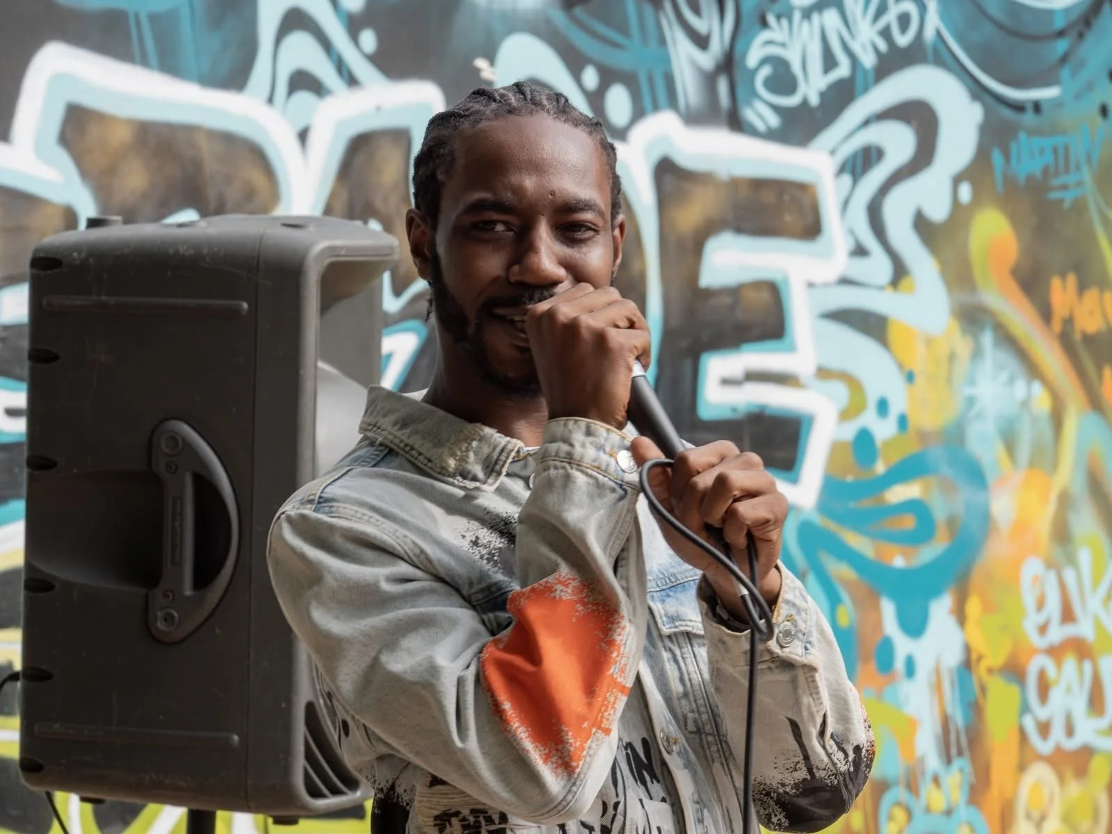 A young man holding a microphone stands in front of a colorful graffiti wall, with a portable speaker by his side, suggesting he is performing or speaking.