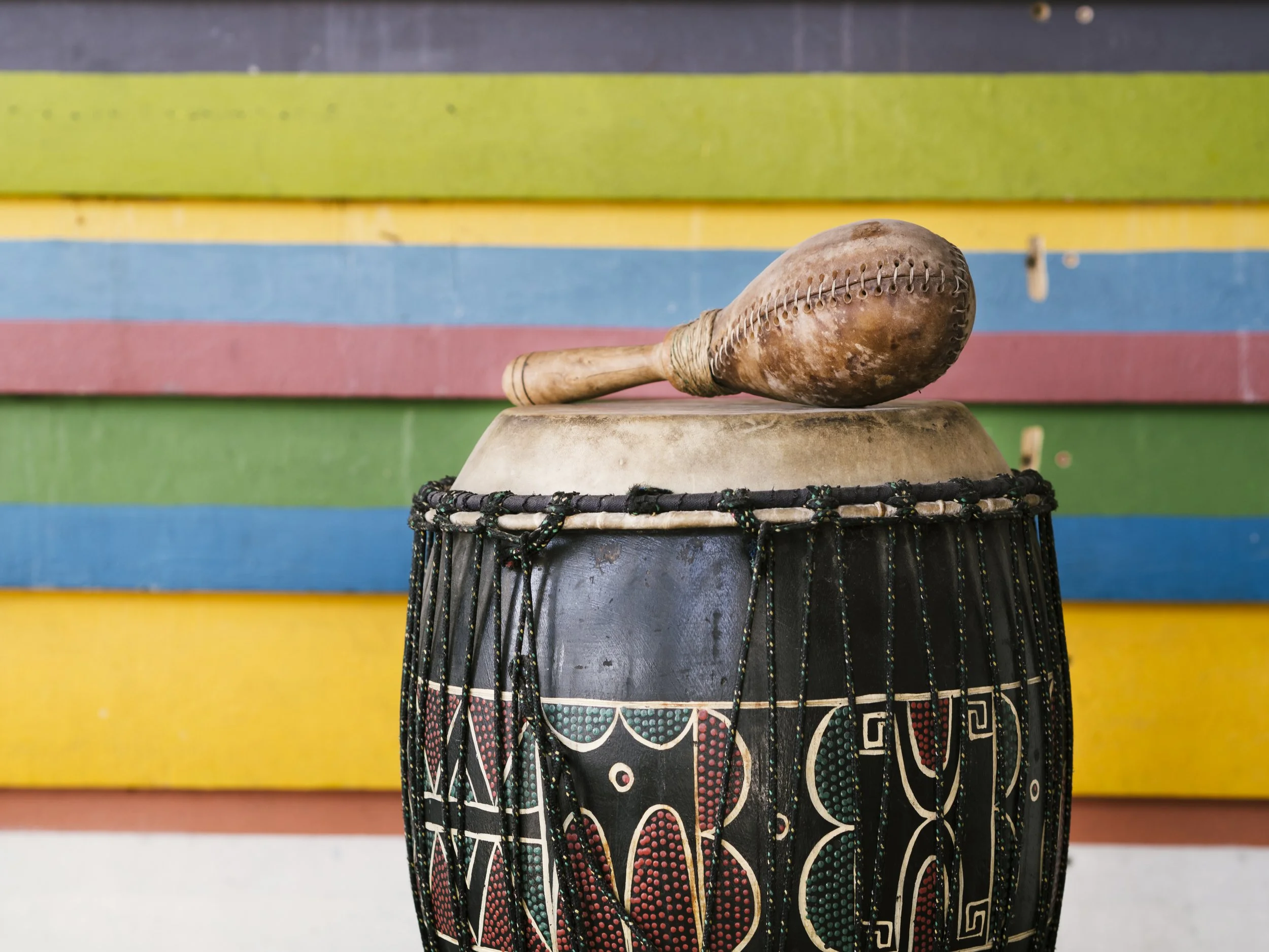 African drum with painted patterns, a wooden maraca, and a colorful wooden background.