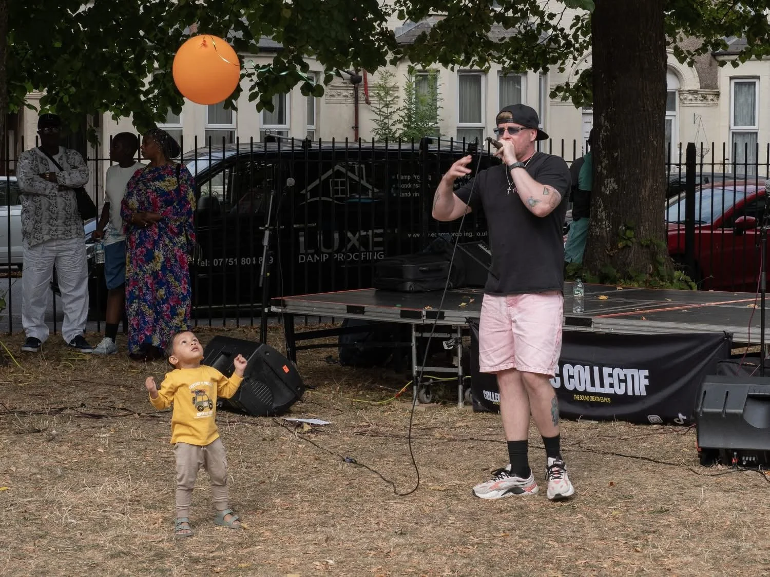 A young boy looking up and raising his fists towards a man singing on a microphone outdoors. The man is wearing sunglasses, a black cap, black t-shirt, pink shorts, and sneakers. The scene is set in a park with a grassy area, trees, and a small stage