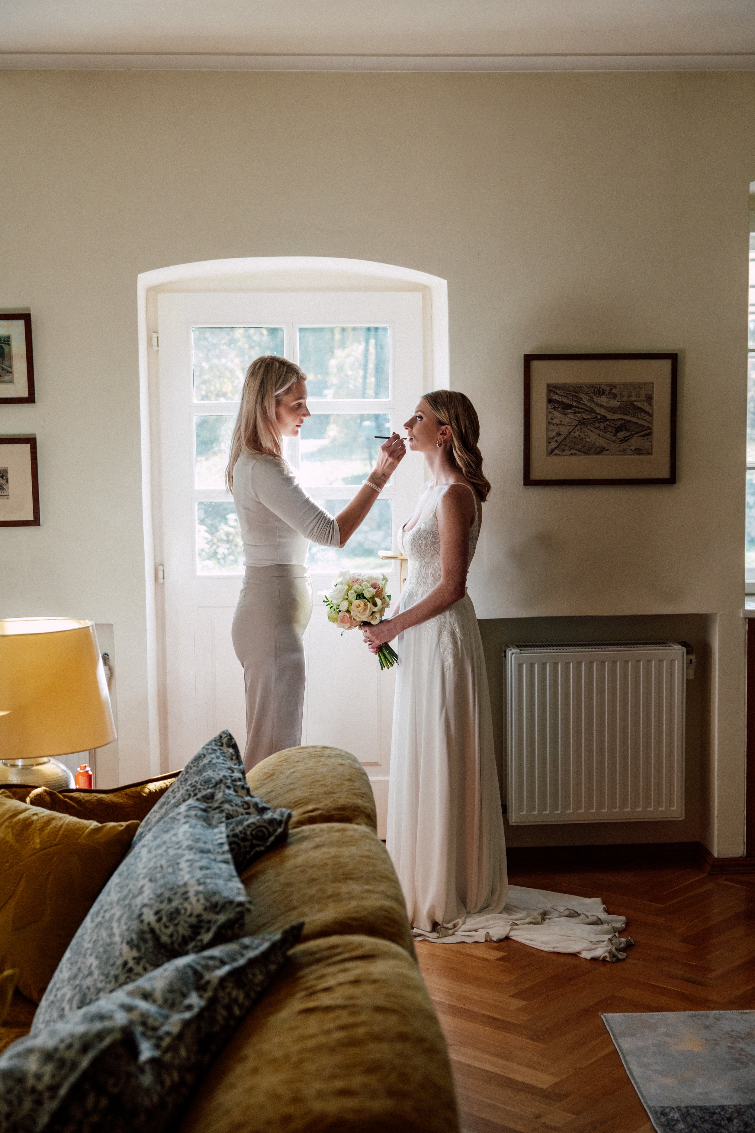 Eine Frau in einem Brautkleid steht in einem Raum und hält einen Blumenstrauß. Eine andere Frau trägt Make-up auf ihr Gesicht auf. Im Hintergrund ist ein Fenster mit Blick ins Grüne zu sehen.