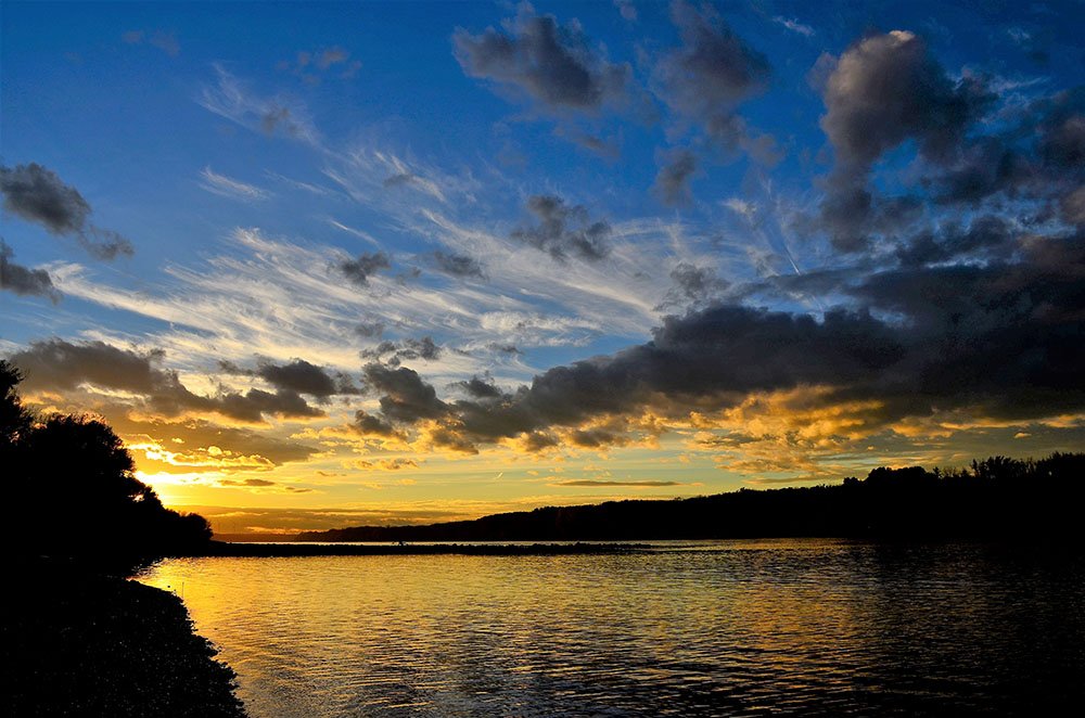 Sonnenuntergang über einem See mit wolkigem Himmel und Bäumen am Ufer.