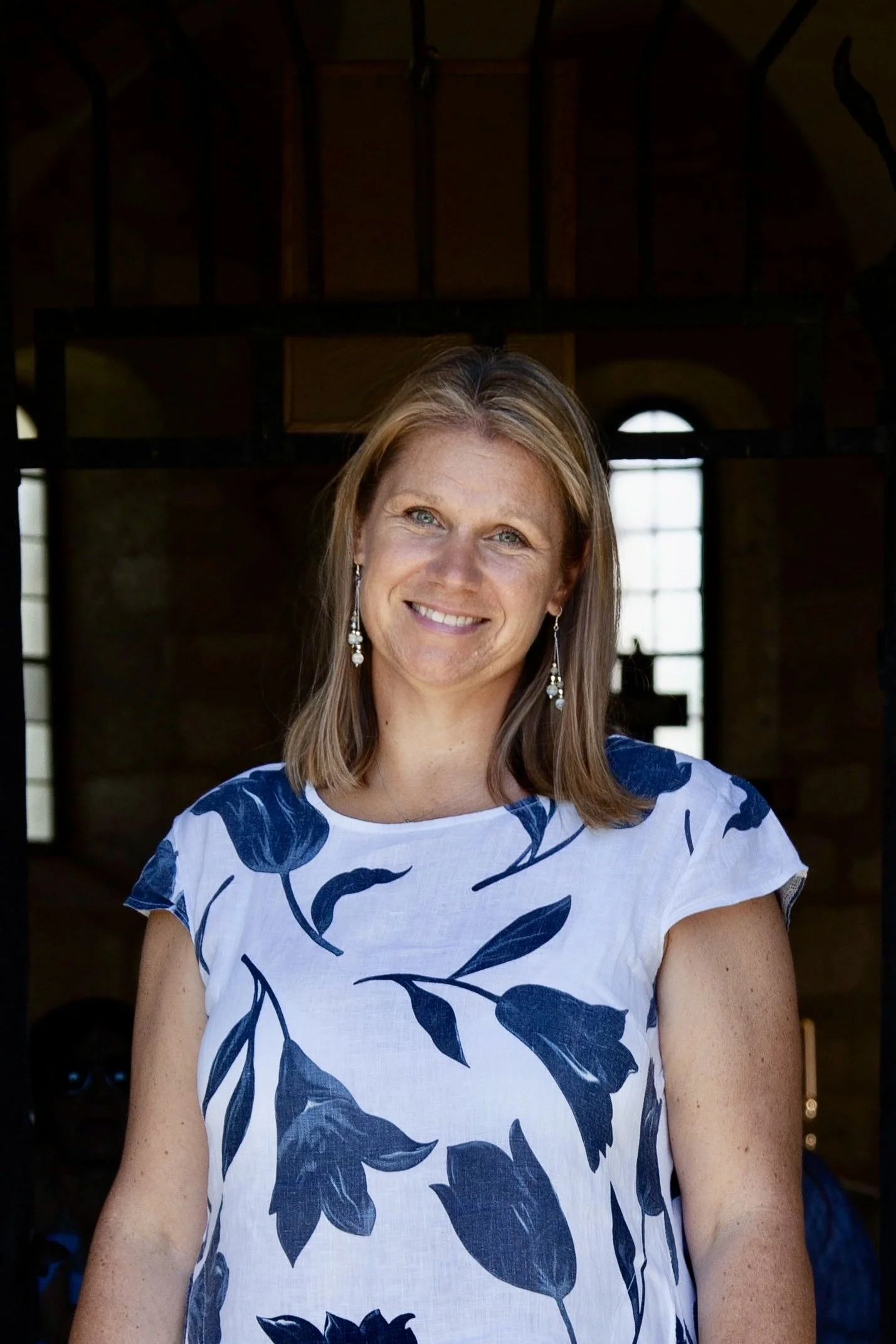 Eine lächelnde Frau mit langem Haar steht vor einer Tafel mit verschiedenen Dokumenten. Sie trägt einen hellen Pullover und einen grauen Schal.