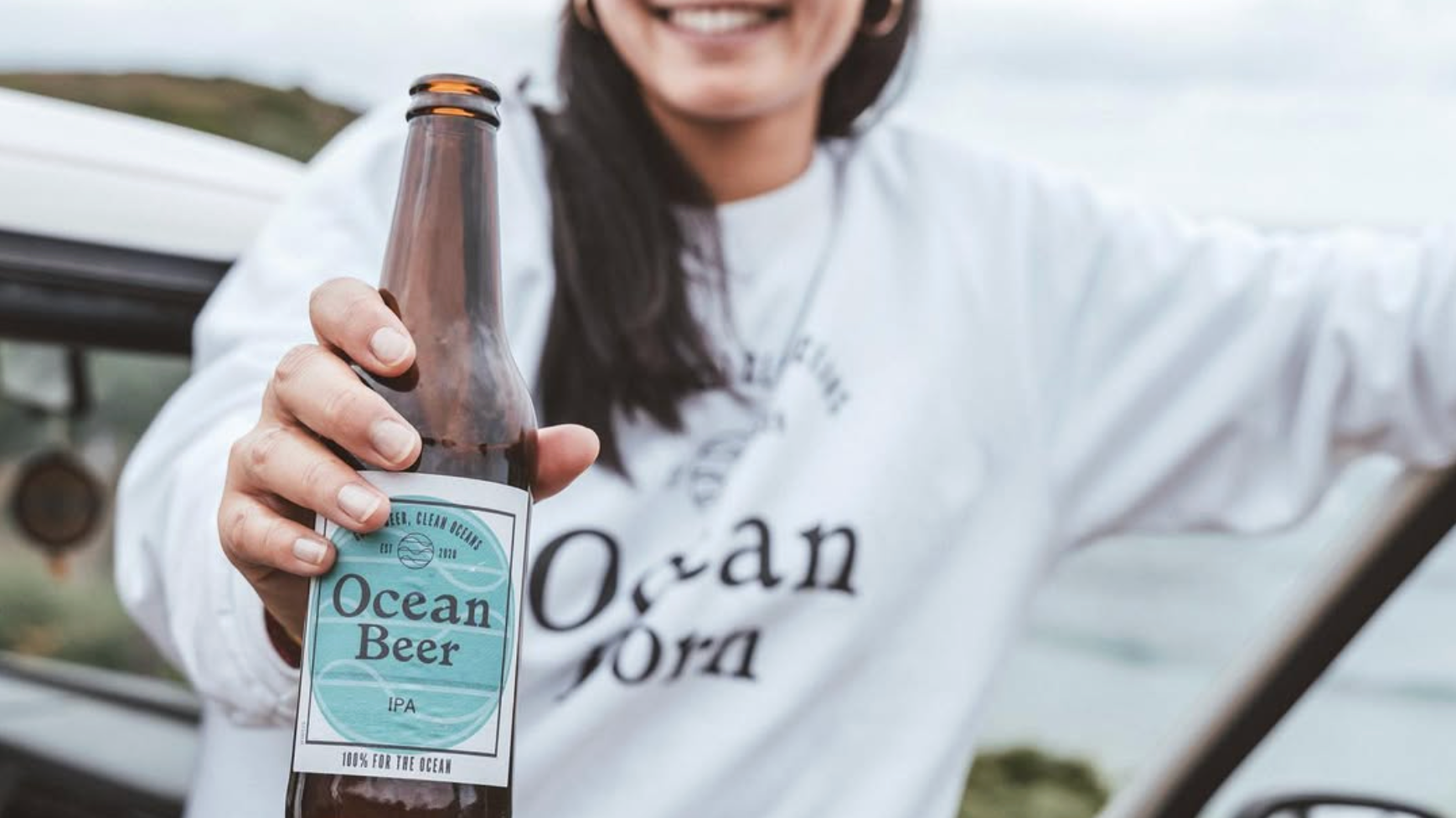 Woman smiling and holding a bottle of Ocean Beer IPA outdoors with a blurred background.