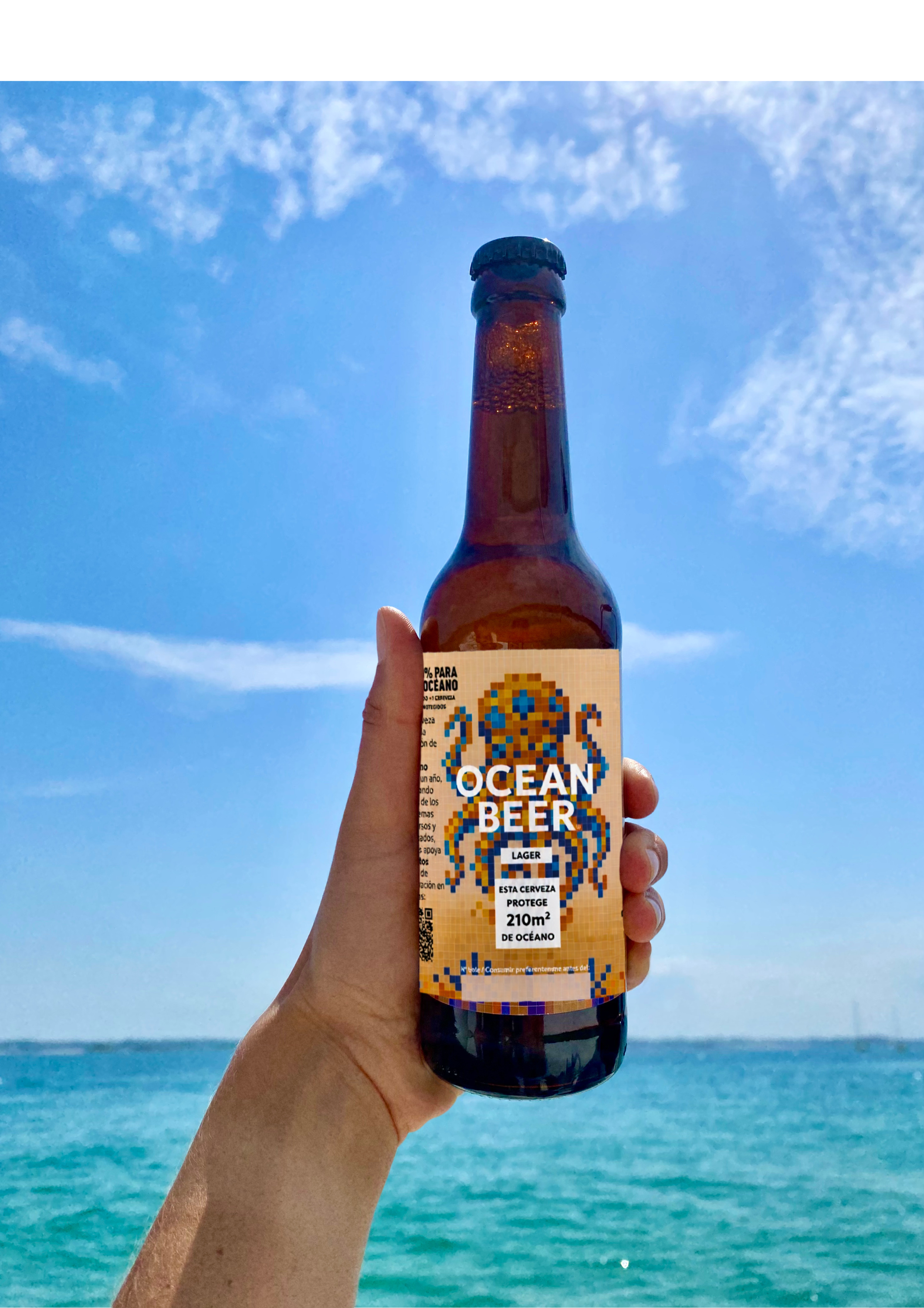 Person holding a bottle of Ocean Beer Lager in focus, with people in the background at a beach or outdoor venue.