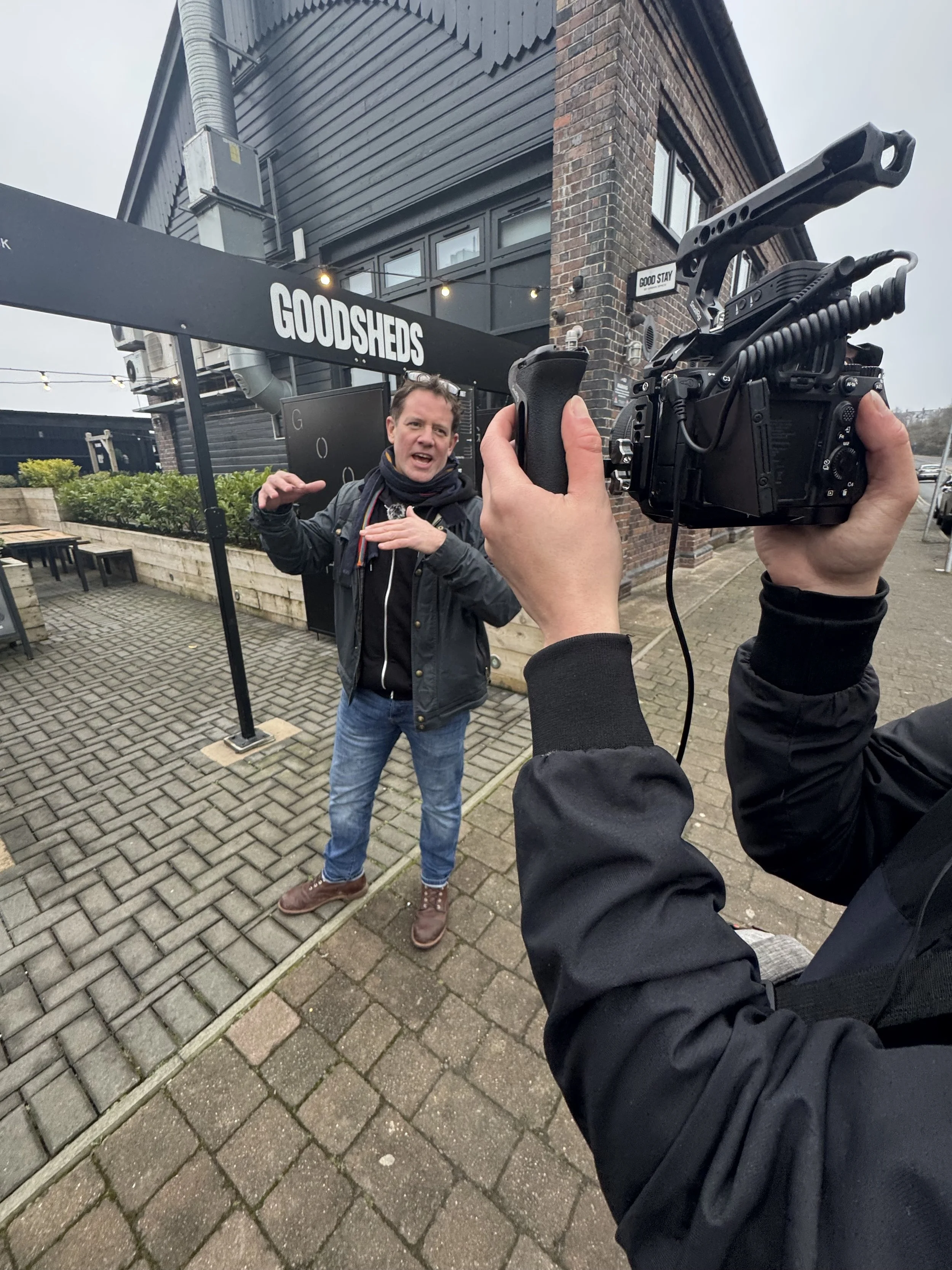 A man is being filmed outside a building labeled 'GOODSHED' by a person holding a video camera. The man appears to be speaking or explaining something, wearing a black jacket, scarf, and jeans. The background shows a sidewalk, some green bushes, and part of a brick building with large windows.