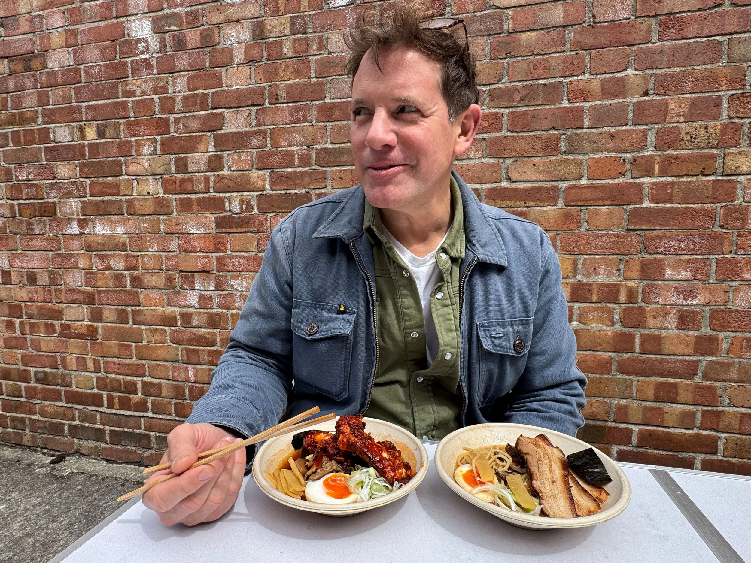 A man sitting at a table with two bowls of ramen noodles, one with soft boiled egg, pork belly, and vegetables, and the other with sliced pork and vegetables, against a brick wall background.