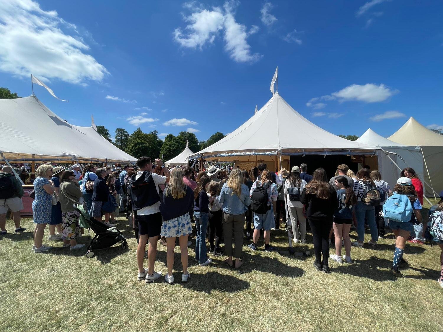 Crowd of people standing in line near white tents at an outdoor event on a sunny day with blue sky and clouds.