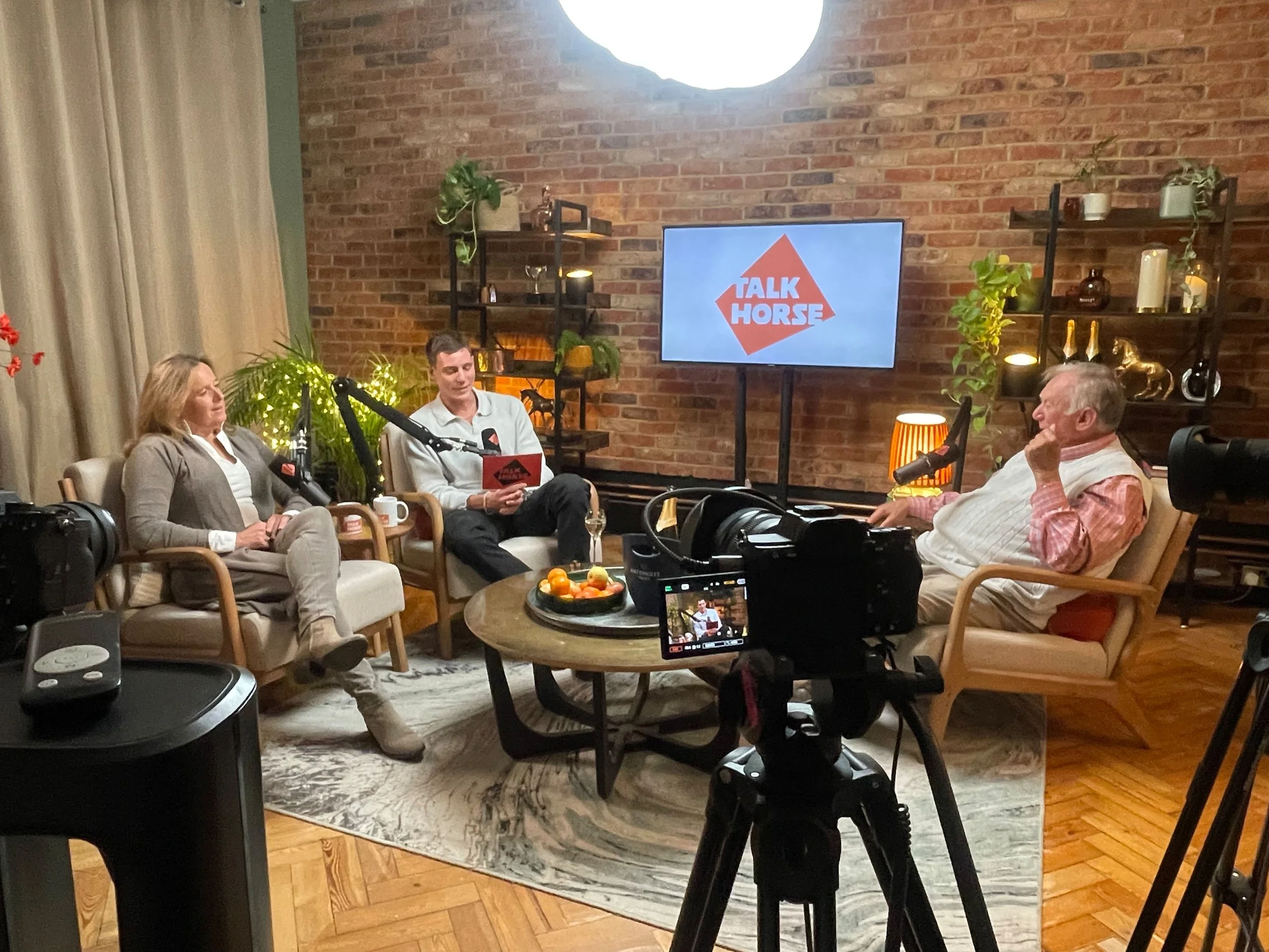 Three people seated in a living room studio with a brick wall background, two of them are speaking into microphones, a camera on a tripod cameras them, a television screen displays the Talk Horse logo, and some decorative plants and items are on shelves.