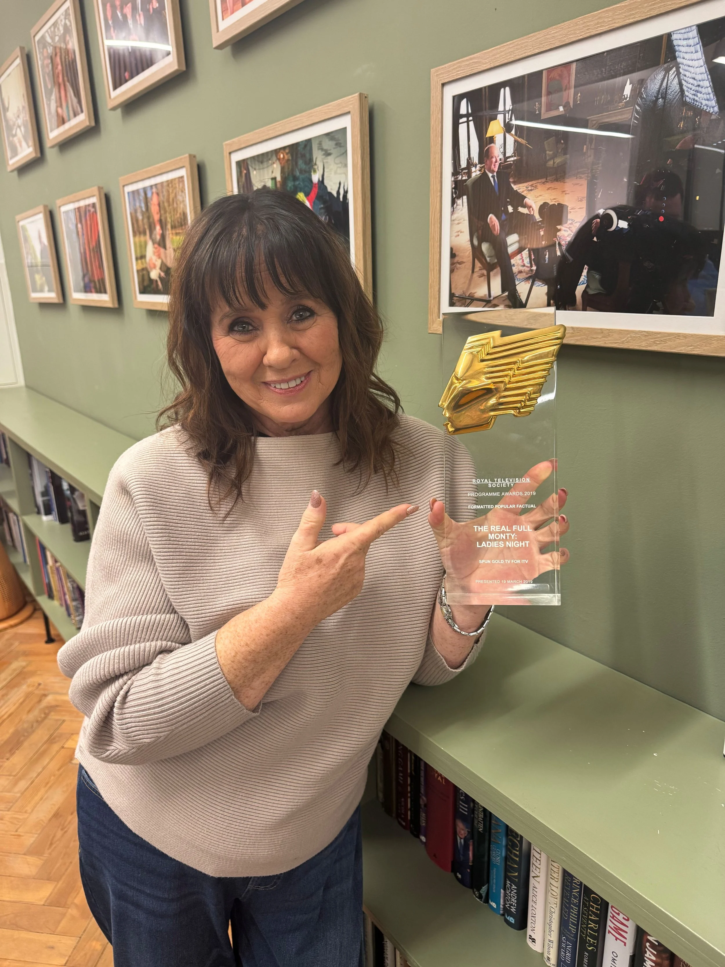 A woman with dark hair and bangs smiling while holding and pointing at a transparent award or plaque with gold accents, standing in a room with a gallery wall of framed photographs and a bookshelf below.