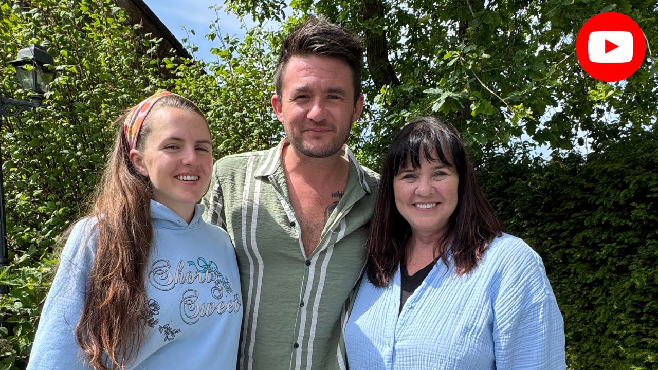 Three people standing outdoors in front of green shrubbery and trees. It is a sunny day. The person in the middle is a man with dark hair and a light green, striped, open-collared shirt. To his left is a woman with dark hair, wearing a light blue shirt, and to his right is a young woman with long brown hair, wearing a light blue hoodie that says 'Short & Sweet' and a colorful headband. All three are smiling.