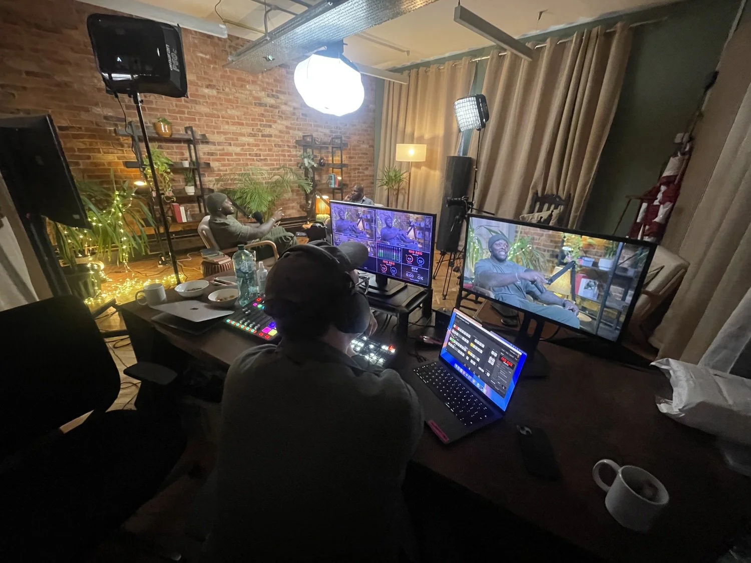A video recording setup with multiple monitors, a person at a mixing console, and a guest on a screen, in a cozy room with brick walls, plants, and warm lighting.