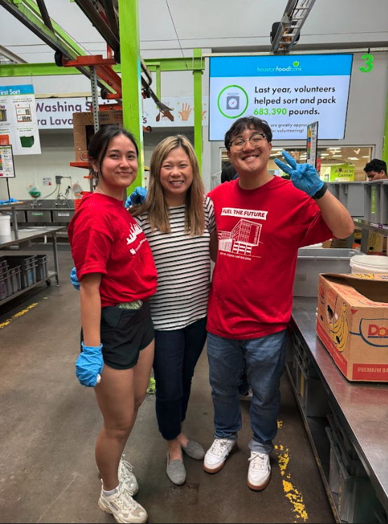 Three volunteers at a food bank, smiling for a photo, with a digital sign in the background displaying a message about the previous year's volunteer efforts.