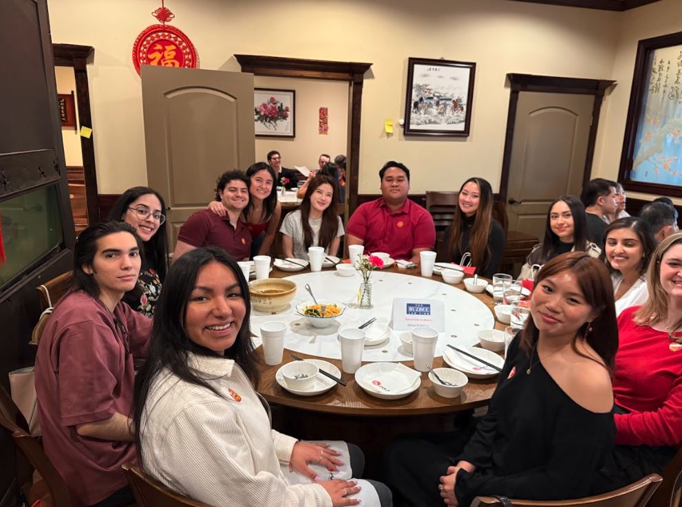 Group of young adults sitting around a round dining table in a restaurant, smiling at the camera, with some empty plates and cups on the table, decorated with a small flower vase and sneeze guards, framed artwork, and Asian-themed decorations in the