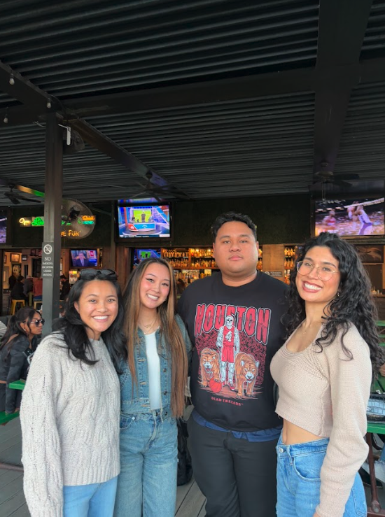 Four friends smiling and posing together in a bar or restaurant with bar shelves and TV screens in the background.