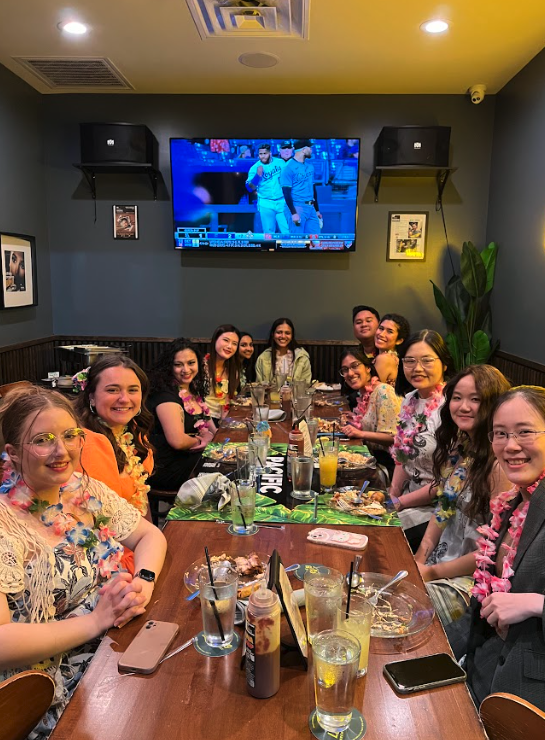 Group of people gathered around a restaurant table enjoying food and drinks, watching a baseball game on a large TV mounted on the wall.