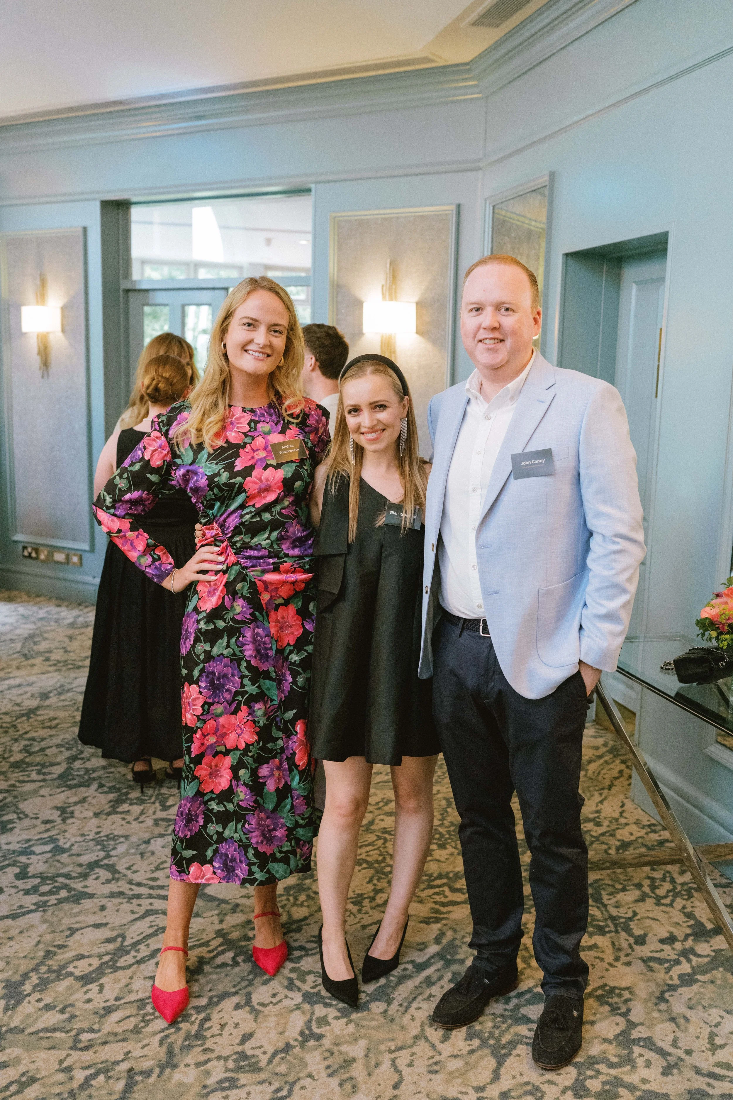 Three adults, two women and one man, smiling and posing together at an indoor event. The woman on the left is wearing a floral dress with red shoes, the woman in the middle is wearing a black dress with black heels, and the man on the right is wearin
