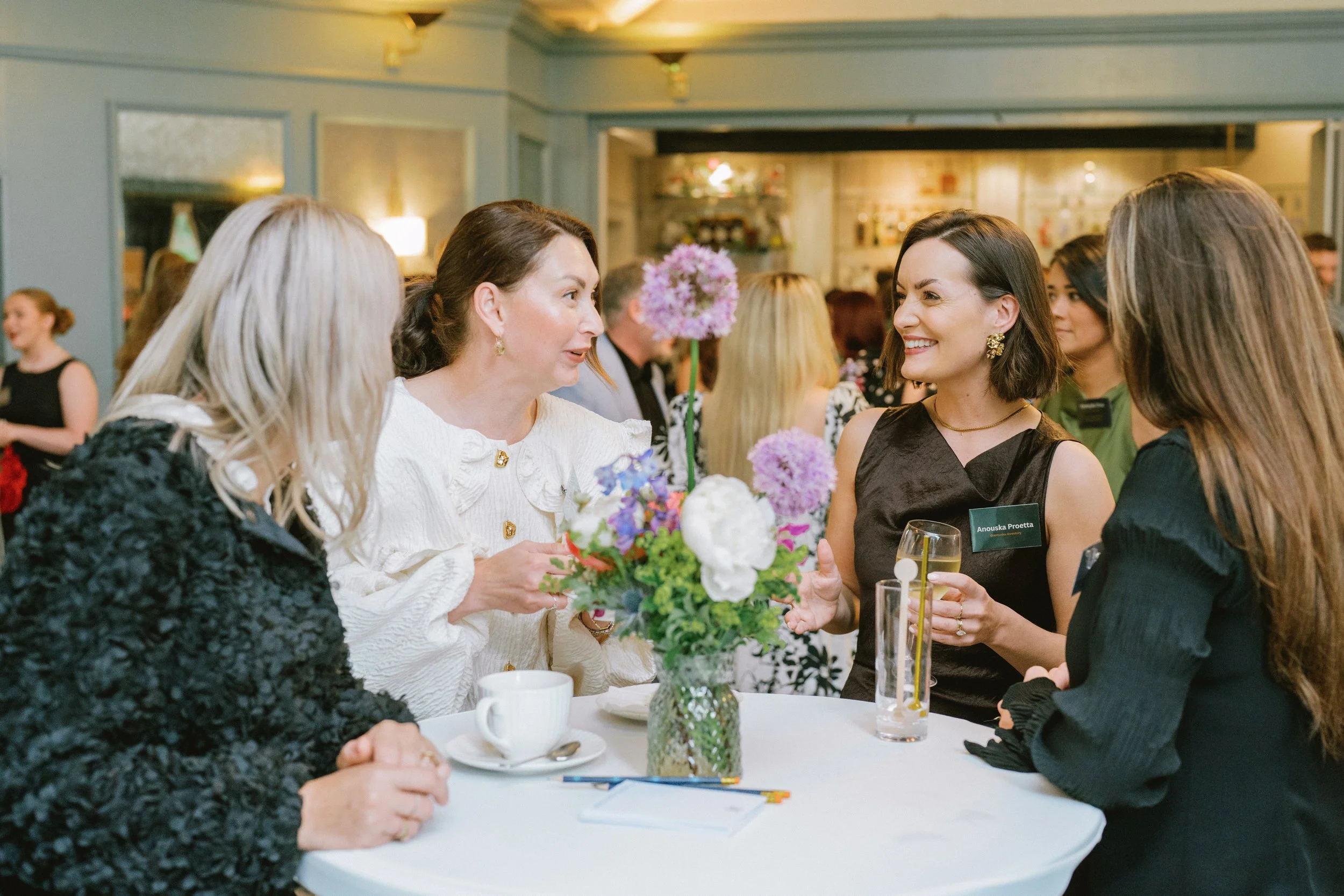 Four women engaged in conversation at a formal event, with a floral centerpiece and drinks on the table.