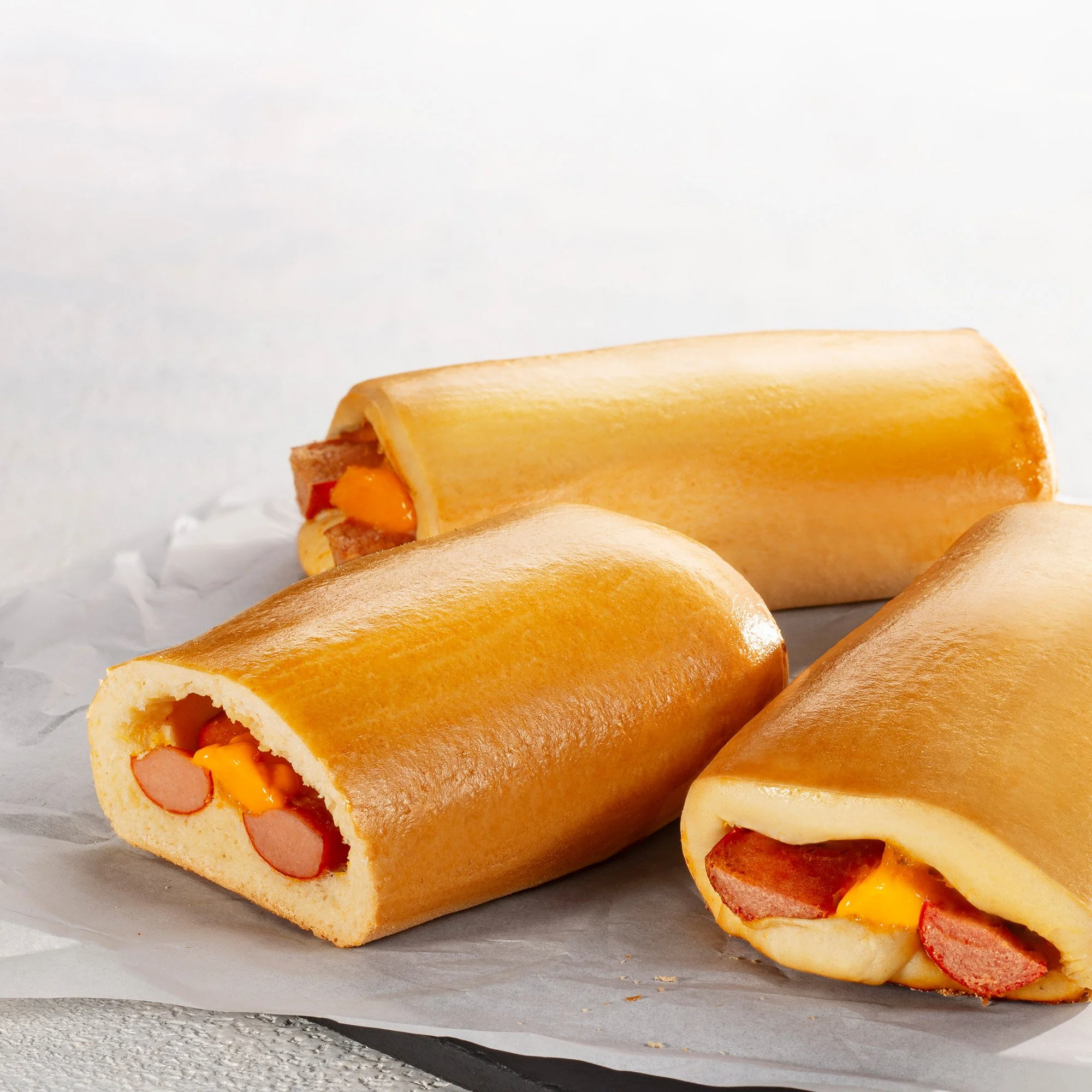 Pães recheados com salsicha e queijo derretido, apresentados sobre papel manteiga.