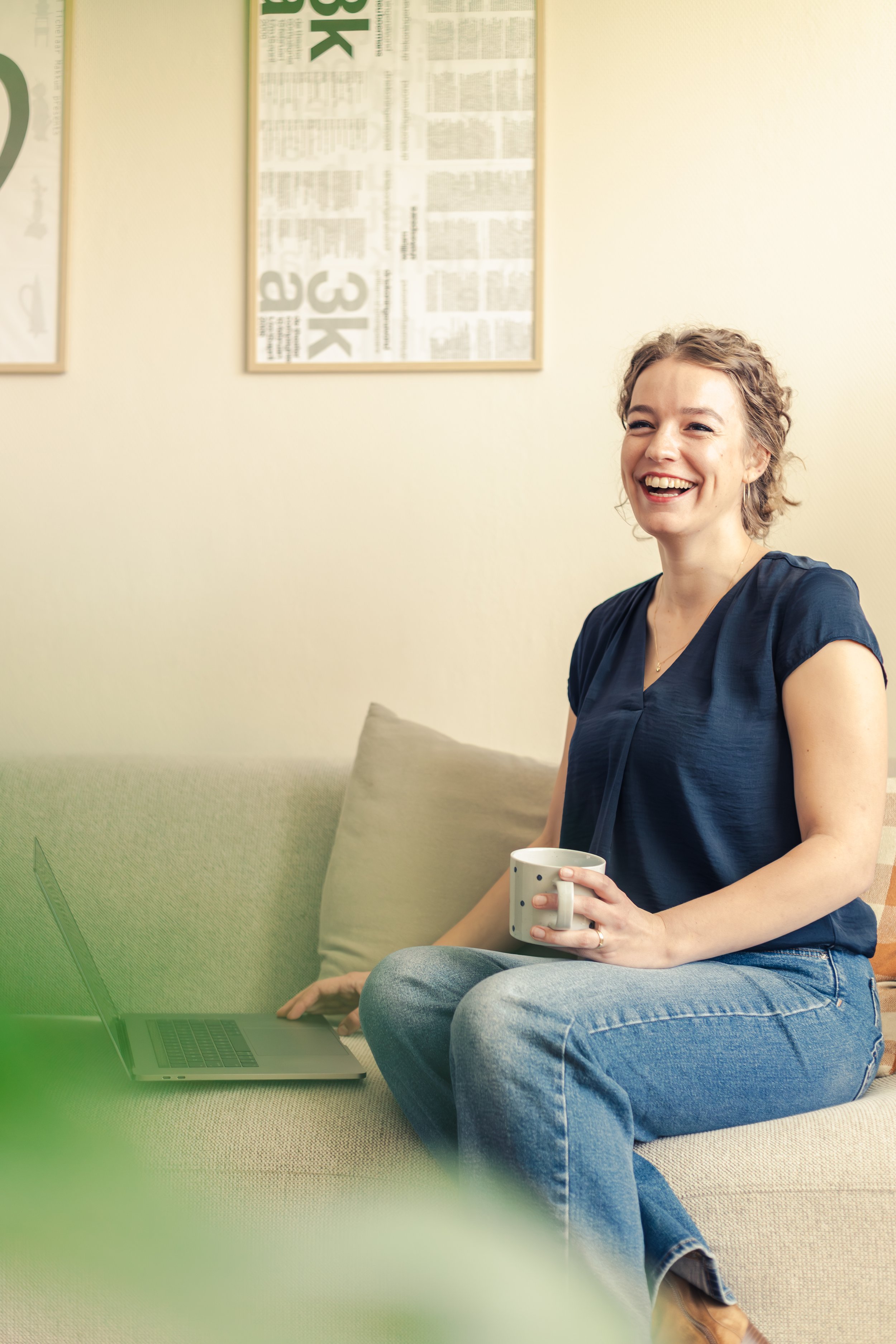A smiling woman sitting on a beige couch, holding a coffee mug, with a laptop open in front of her, in a room with wall posters in the background.