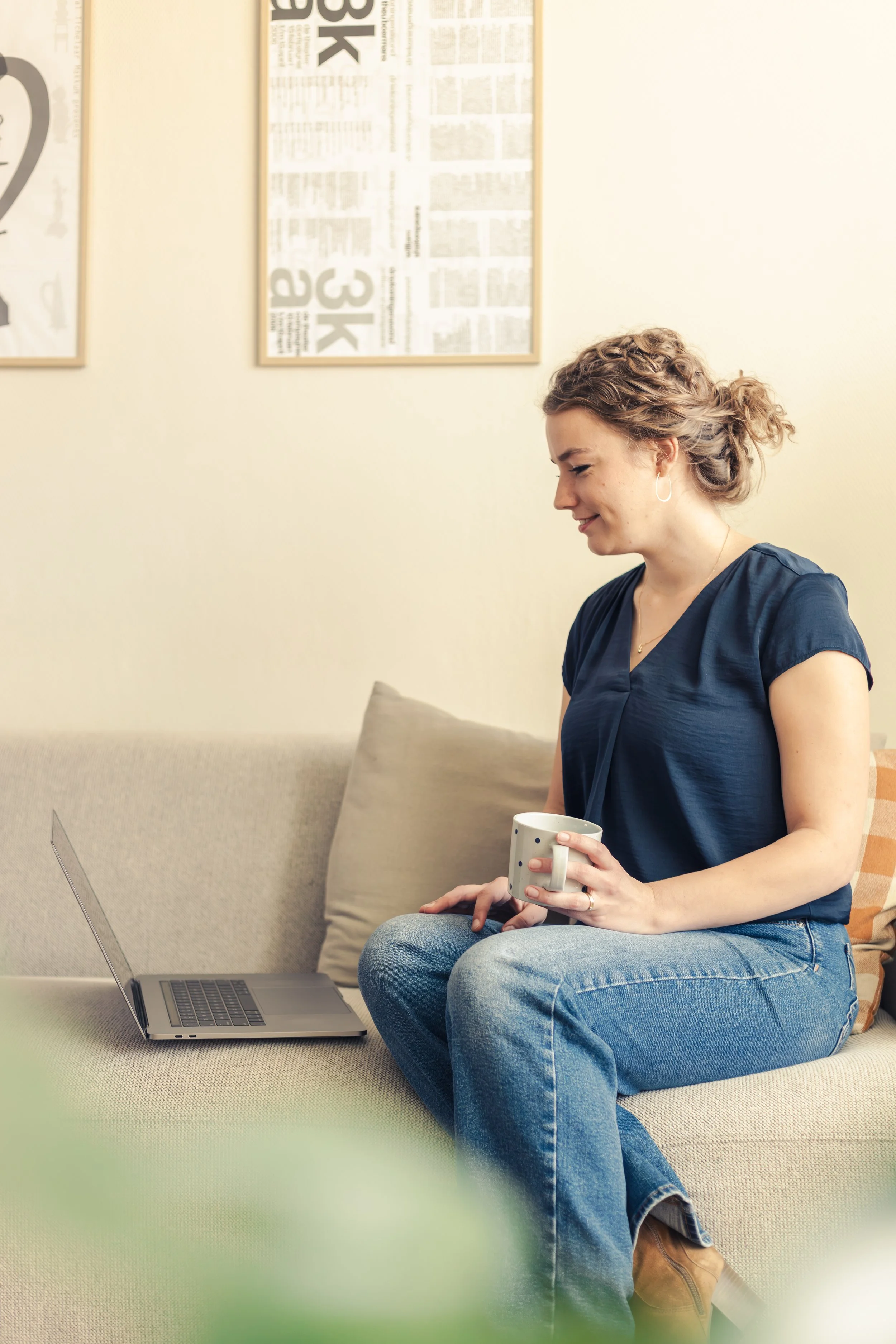 A woman with curly hair sitting on a beige sofa, holding a mug, with a laptop open in front of her. She is wearing a dark blue top and blue jeans, smiling and looking down. There are framed newspaper clippings on the wall behind her, and a pillow on the sofa.
