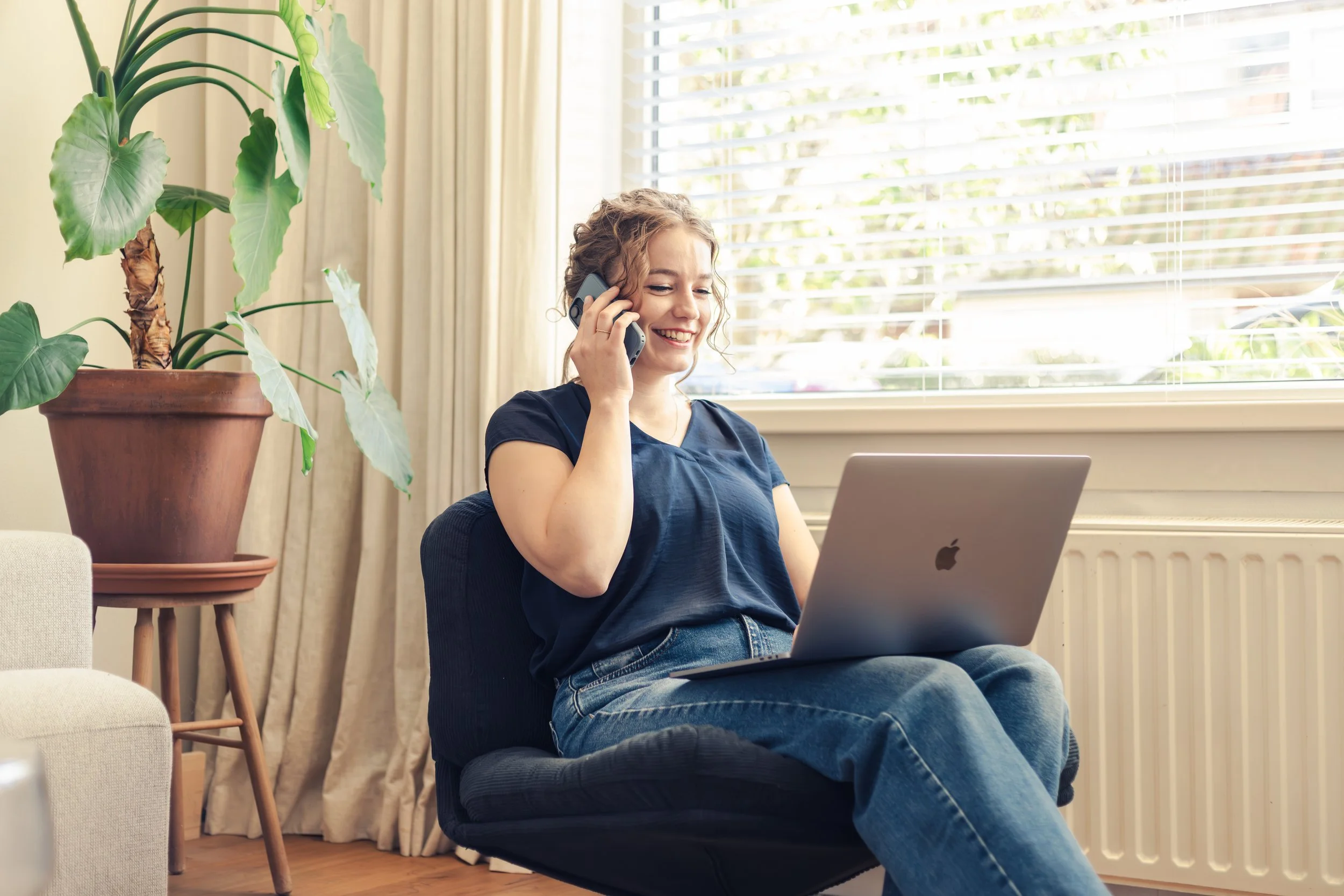 Amy, een vrouw met krullend haar zit op een stoel en praat via een telefoon, terwijl ze een laptop op schoot heeft. Ze bevindt zich in een lichte ruimte met grote raamjaloezieën, een grote kamerplant in een pot en lichtgekleurde gordijnen.