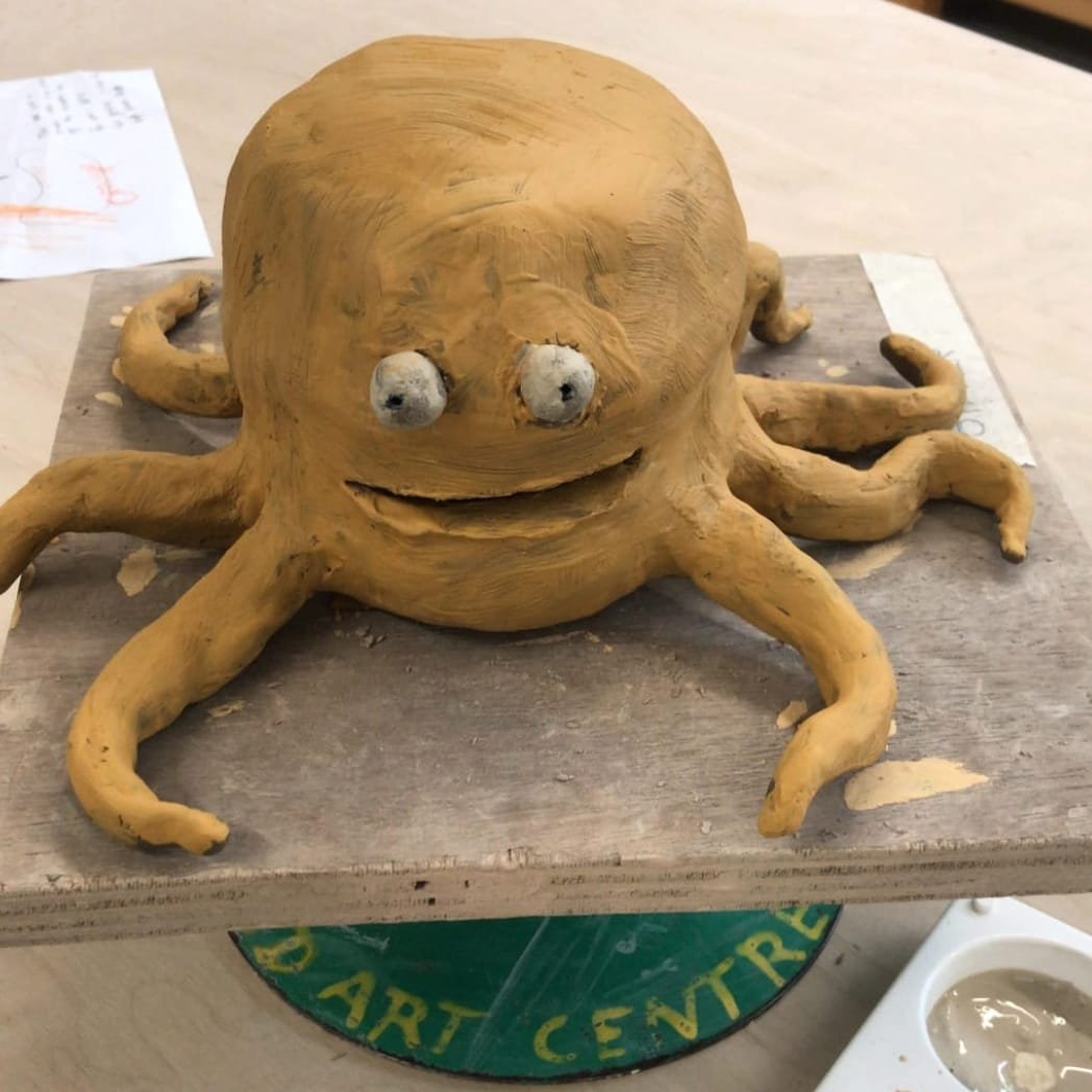 Clay octopus sculpture with a smiling face and googly eyes on a wooden board.
