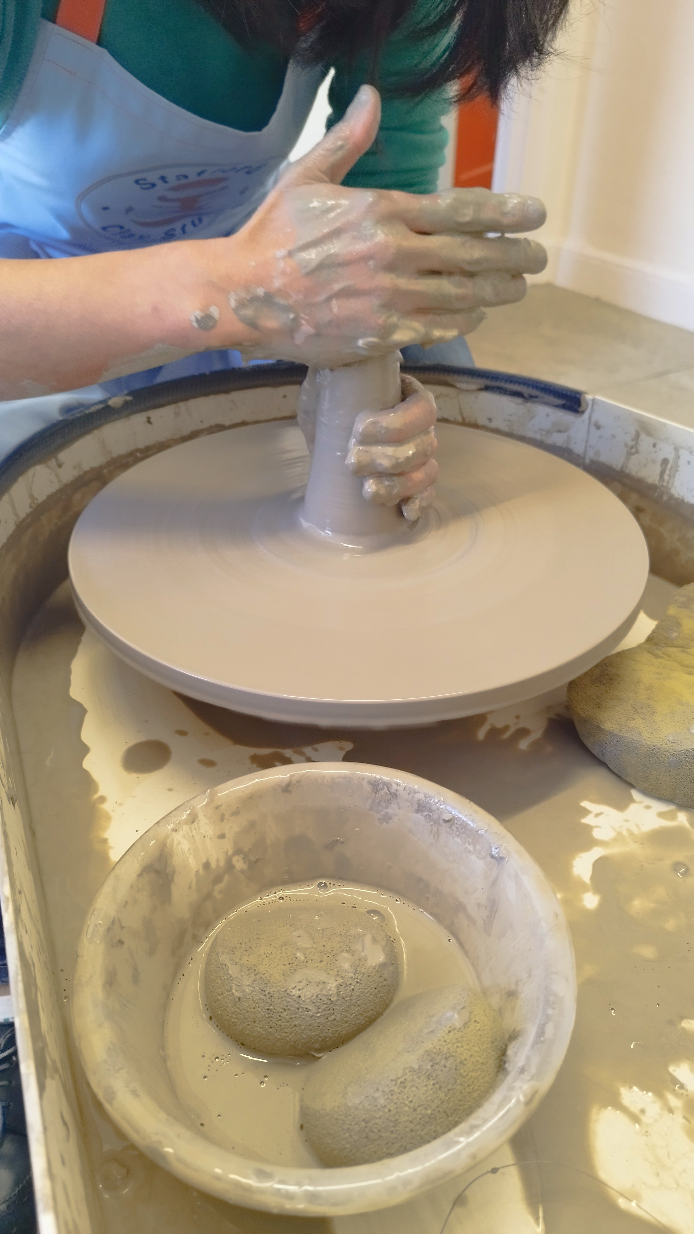 Person shaping clay on a pottery wheel, with hands covered in wet clay and sponges nearby.
