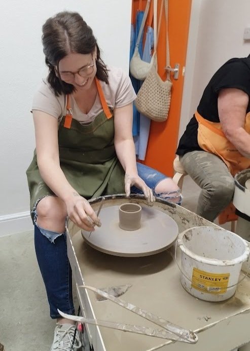 Person shaping clay on a pottery wheel, wearing a green apron and glasses, focused on creating a pot. A bucket of water and pottery tools are nearby.