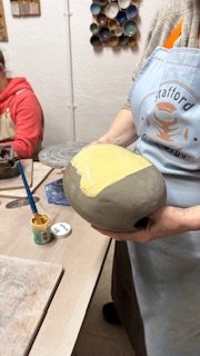 Person applying yellow glaze to a pottery piece in a ceramics studio, with paintbrush and tools on the table.
