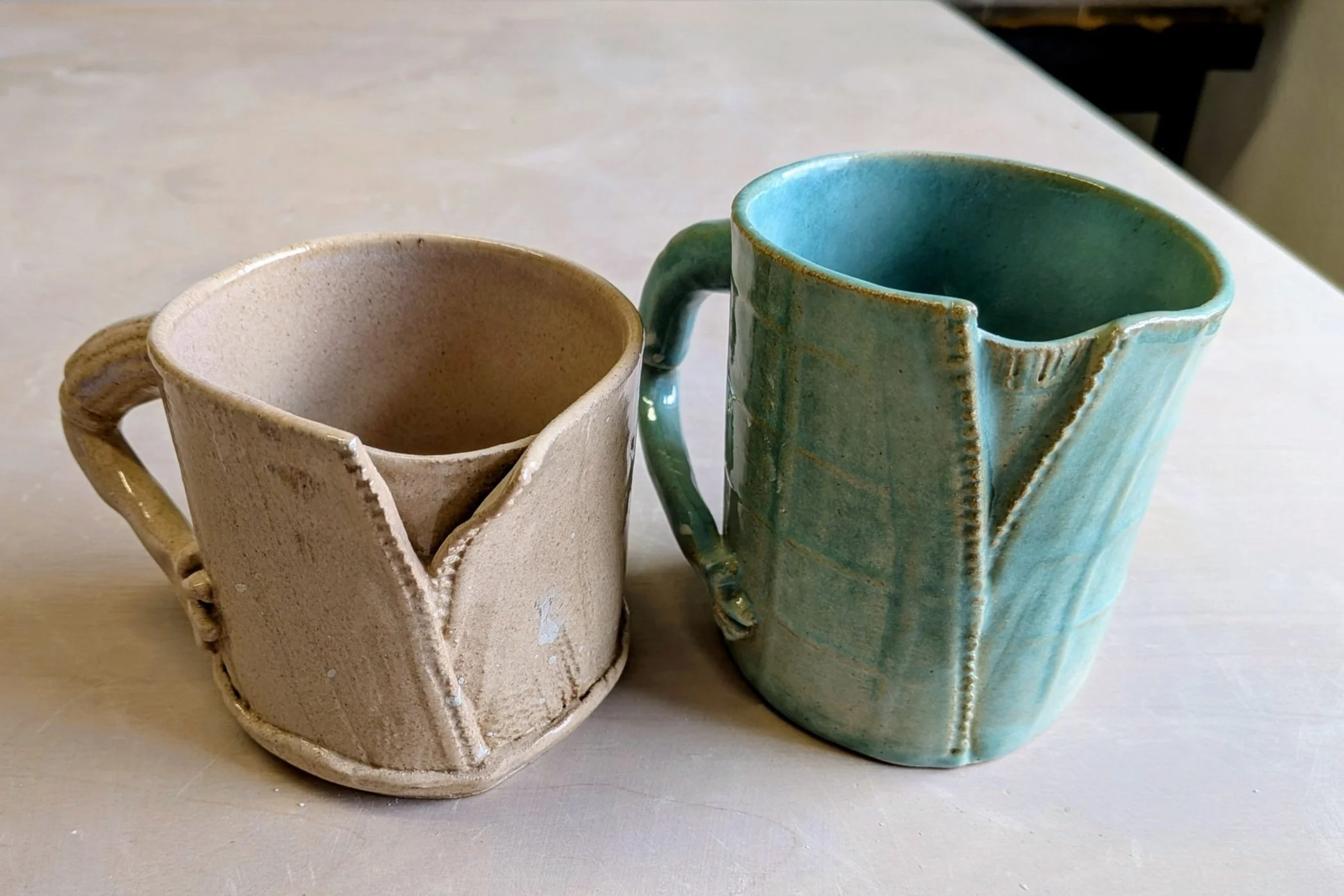Two uniquely designed ceramic mugs with sculpted cloth-like textures, one beige and one blue, placed on a table.