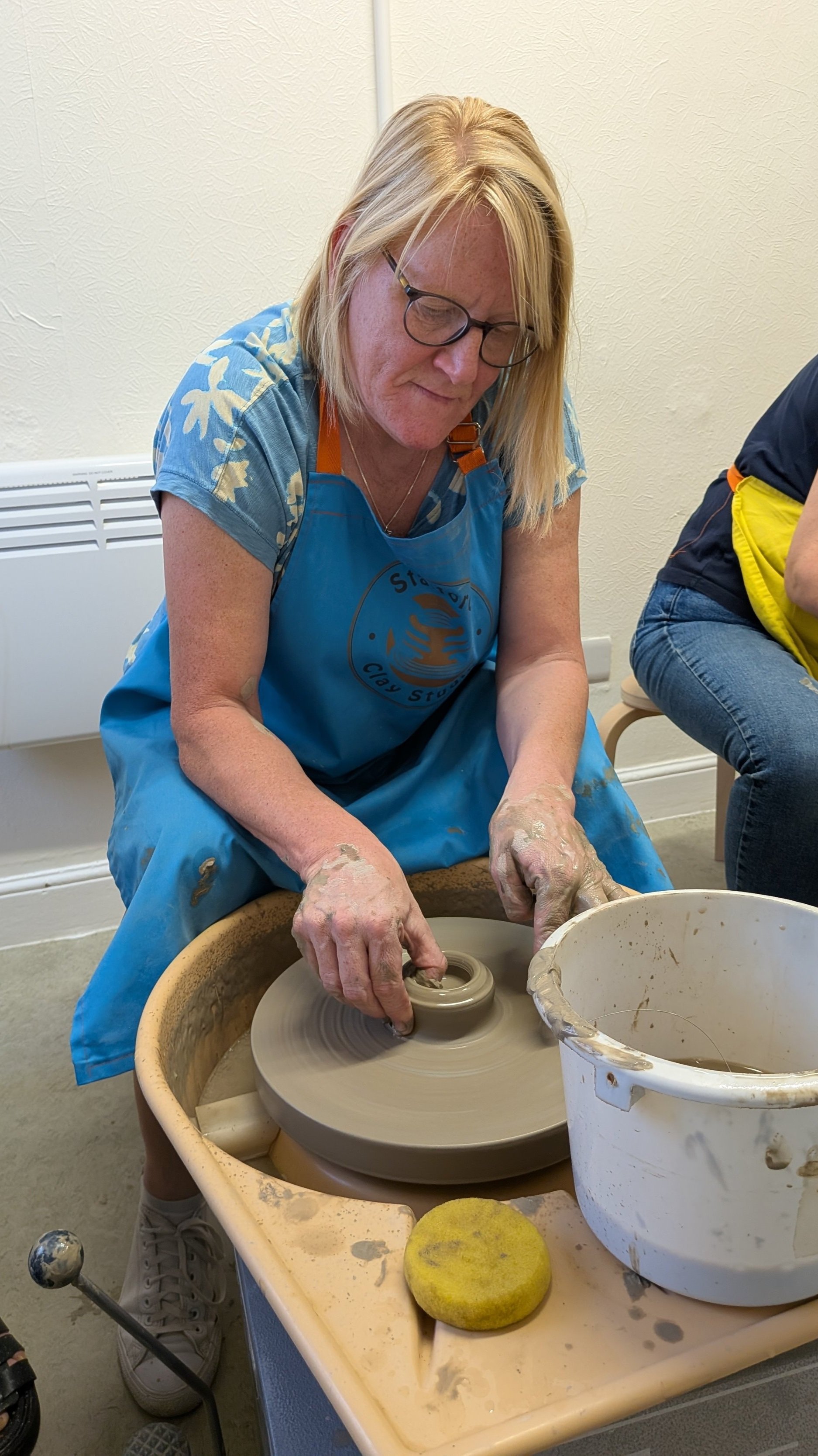 Person working with clay on a pottery wheel, wearing a blue apron and glasses, with hands shaping the clay. A yellow sponge and a bucket are nearby.