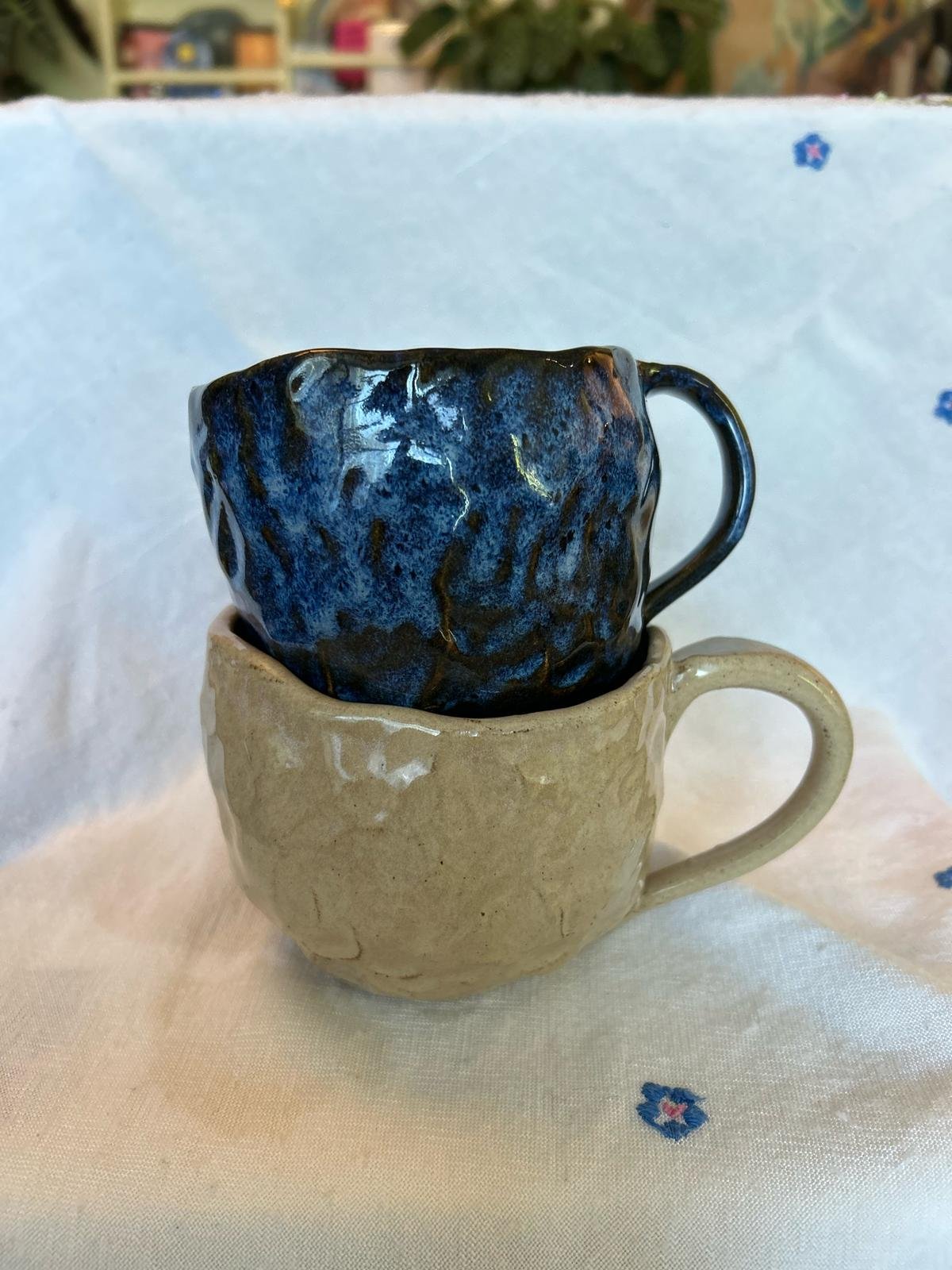 Two handmade ceramic mugs stacked, one blue with dark texture, the other beige, on a white cloth with small blue floral designs.