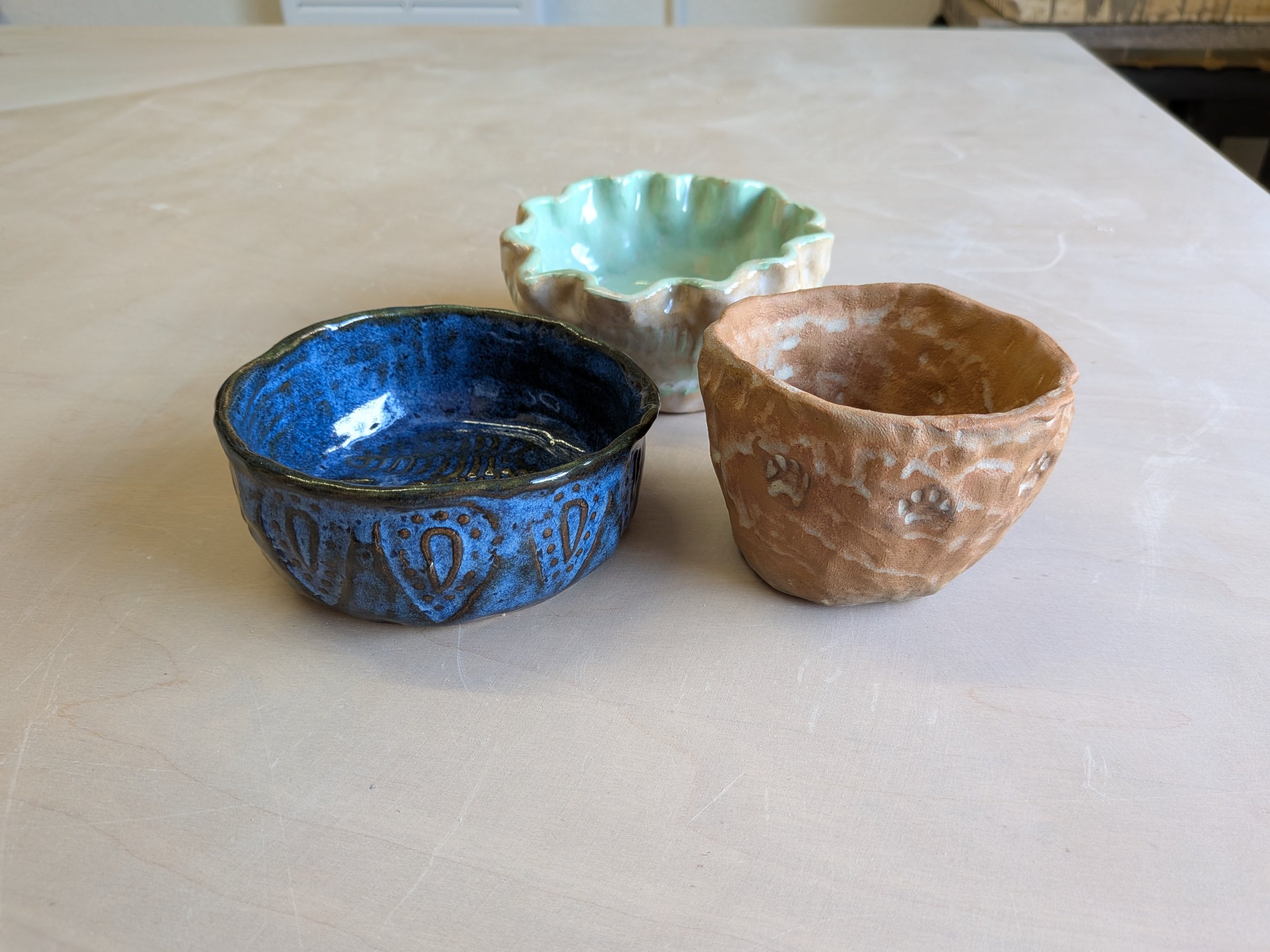 Three hand-crafted ceramic bowls, one blue with patterns, one light green with a wavy edge, and one textured brown, arranged on a light surface.