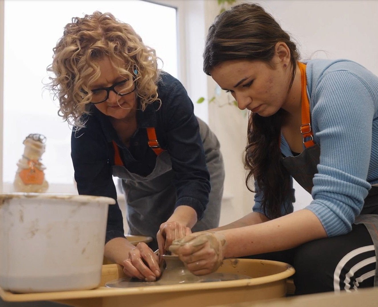 Two women working on a pottery wheel, focusing on shaping clay. They are wearing aprons and appear to be in a pottery studio.